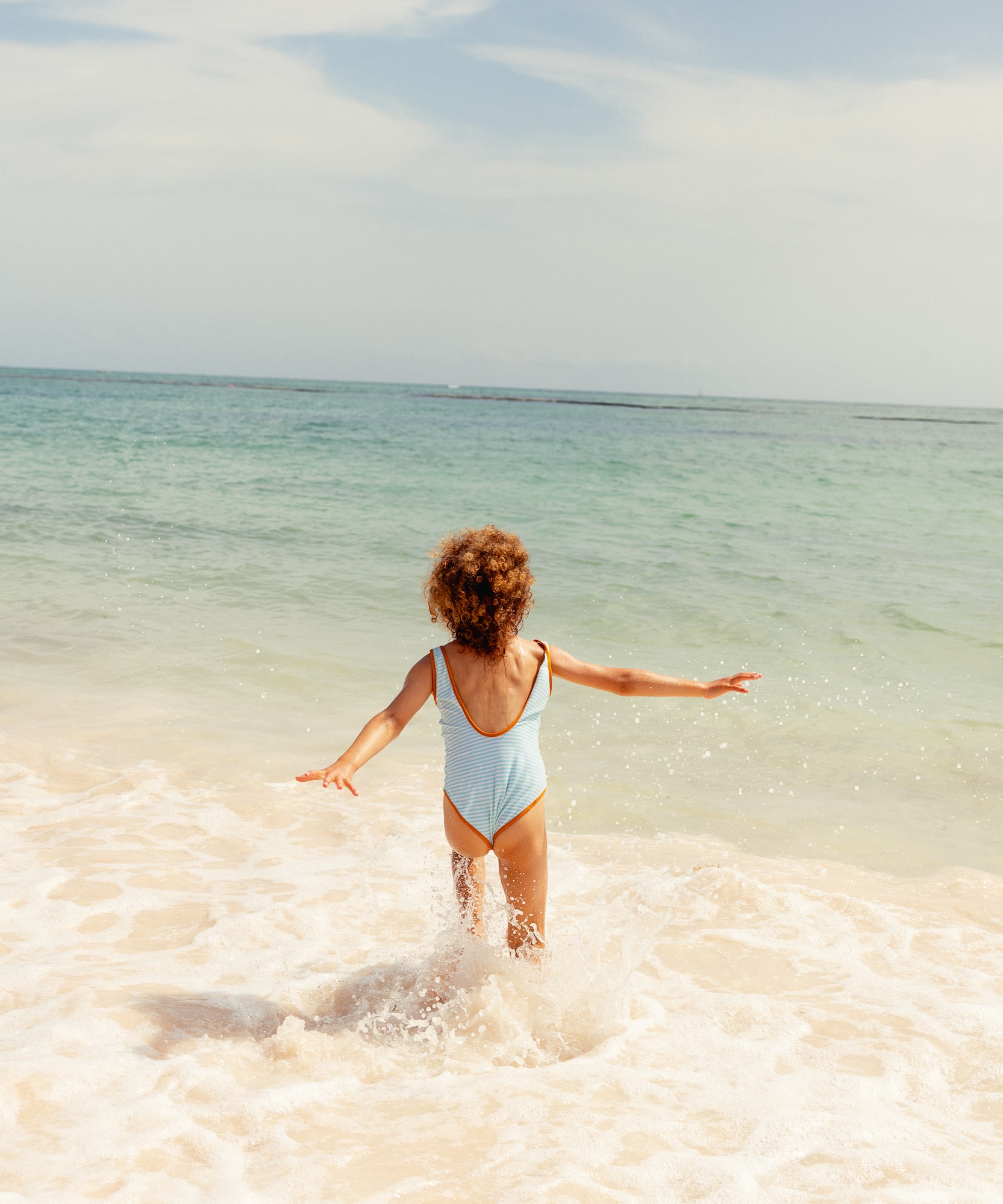 Child in blue striped swimsuit splashes in ocean waves at sunny beach.