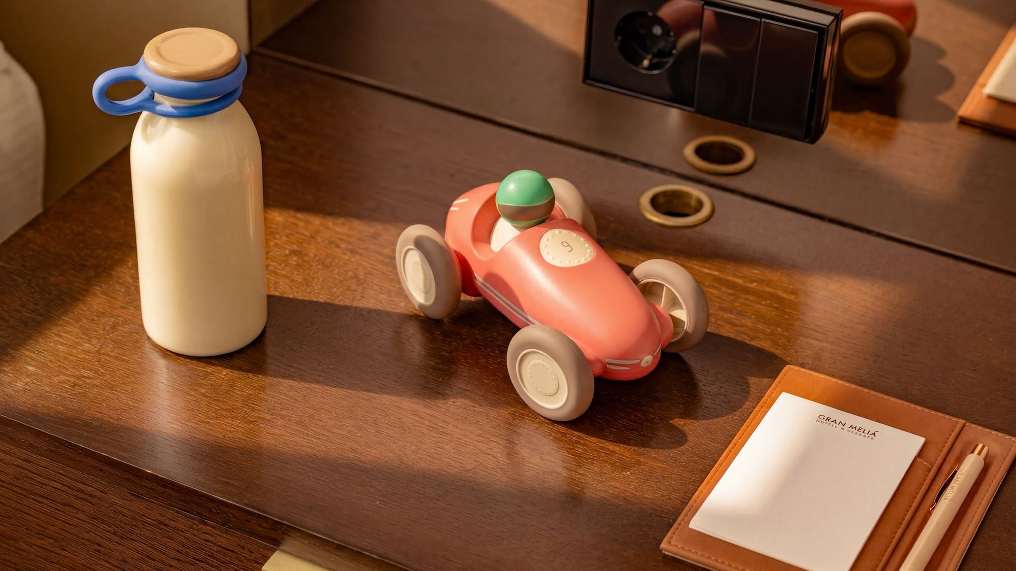 Pink toy race car, bottle, and Gran Meliá notepad on a wooden hotel desk.