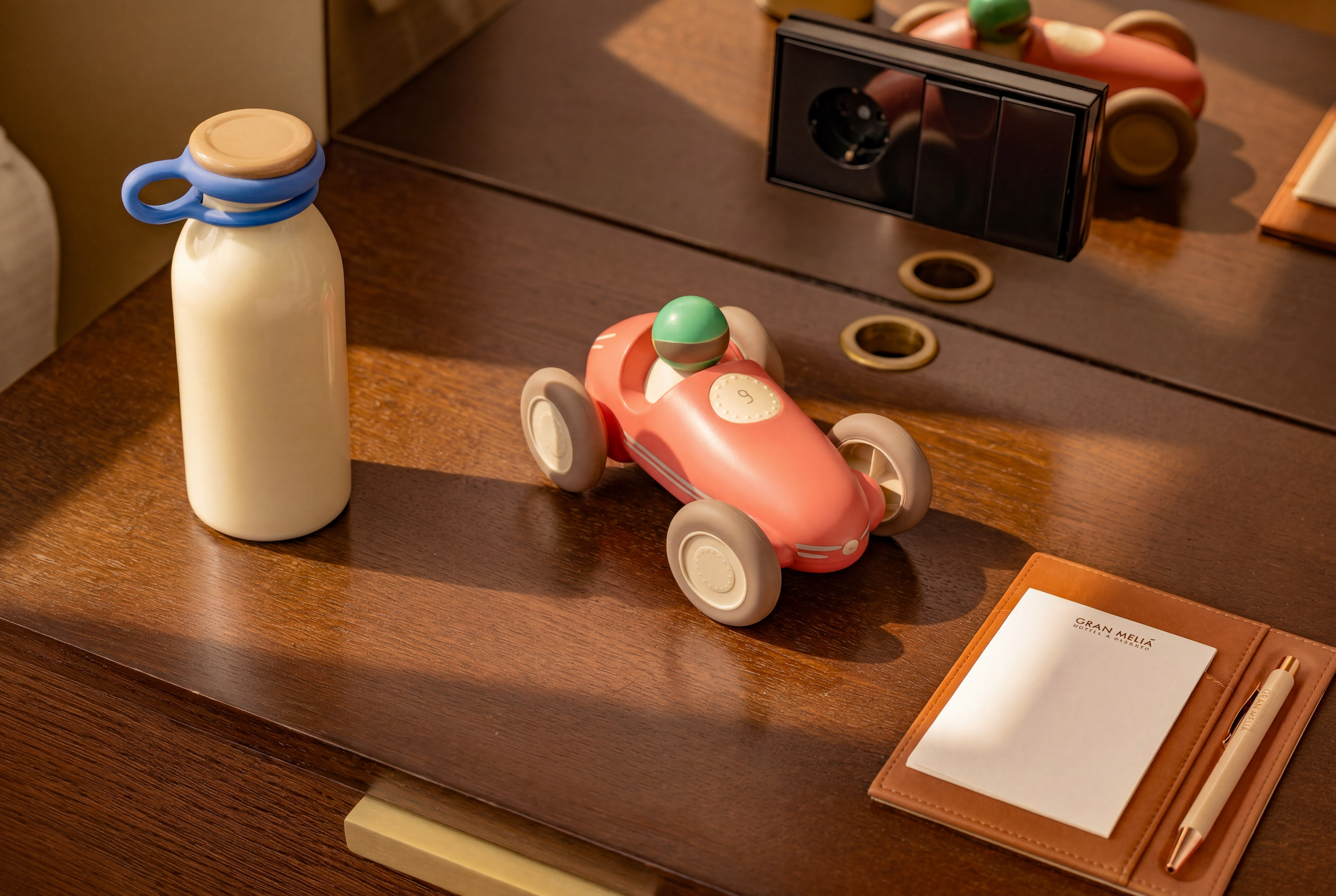 Pink toy race car, bottle, and Gran Meliá notepad on a wooden hotel desk.