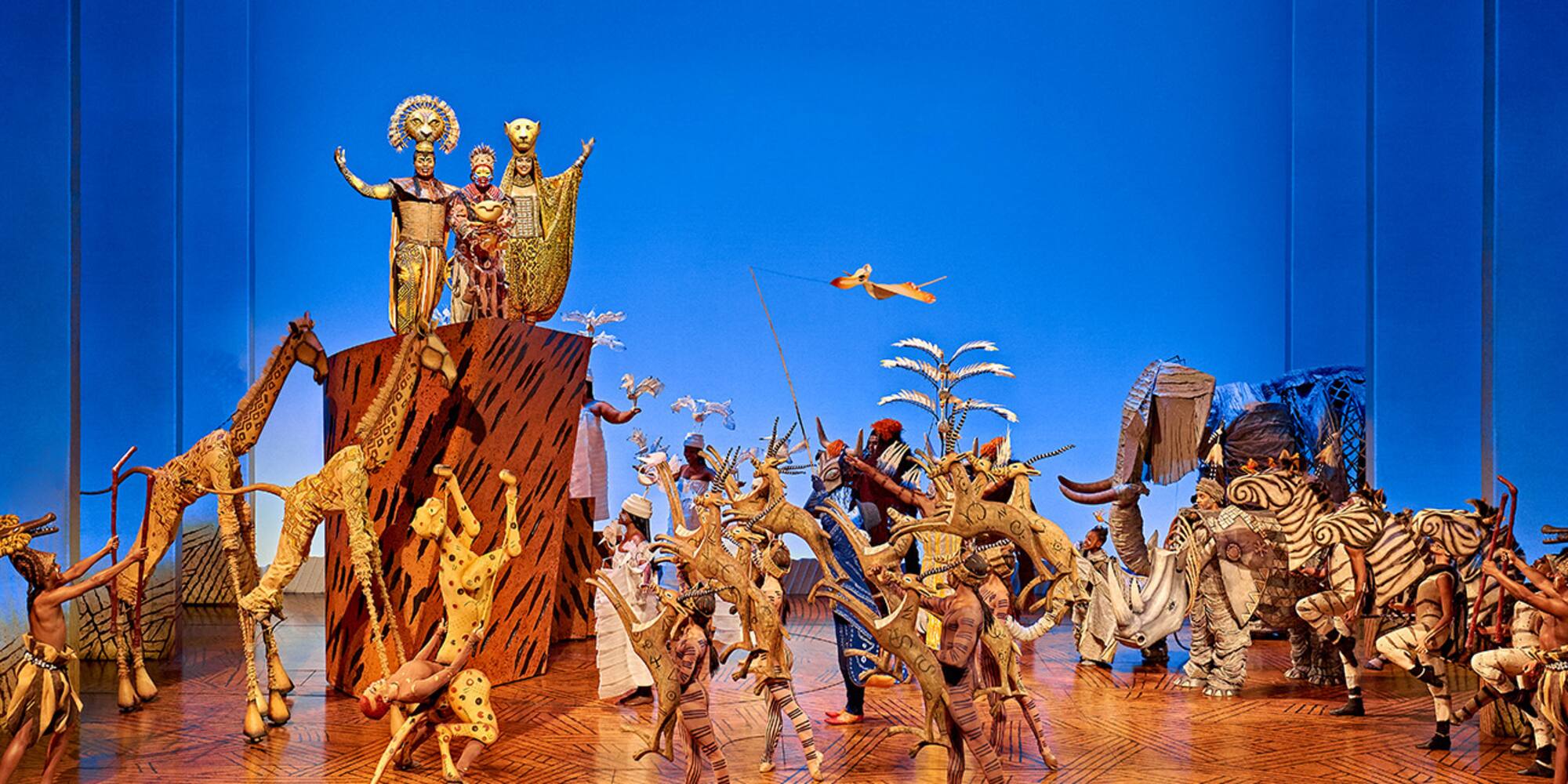 Lion King musical stage: Rafiki presenting Simba, elaborate animal costumes and puppets.