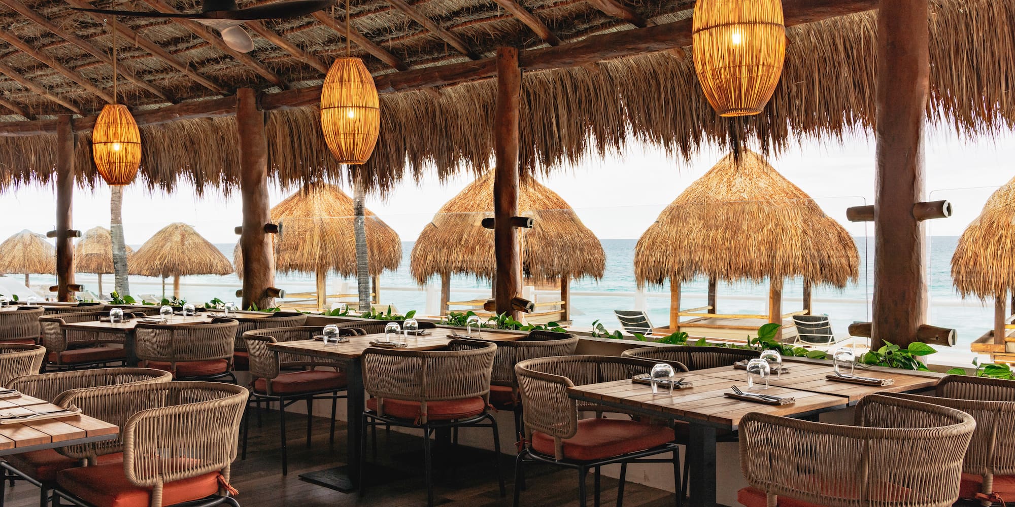 Beachfront palapa restaurant with ocean view, woven lamps, and set dining tables.