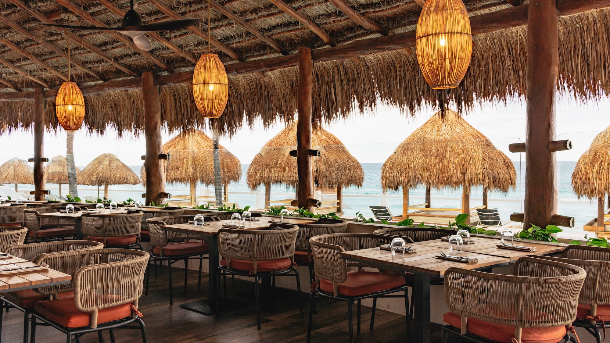 Beachfront palapa restaurant with ocean view, woven lamps, and set dining tables.