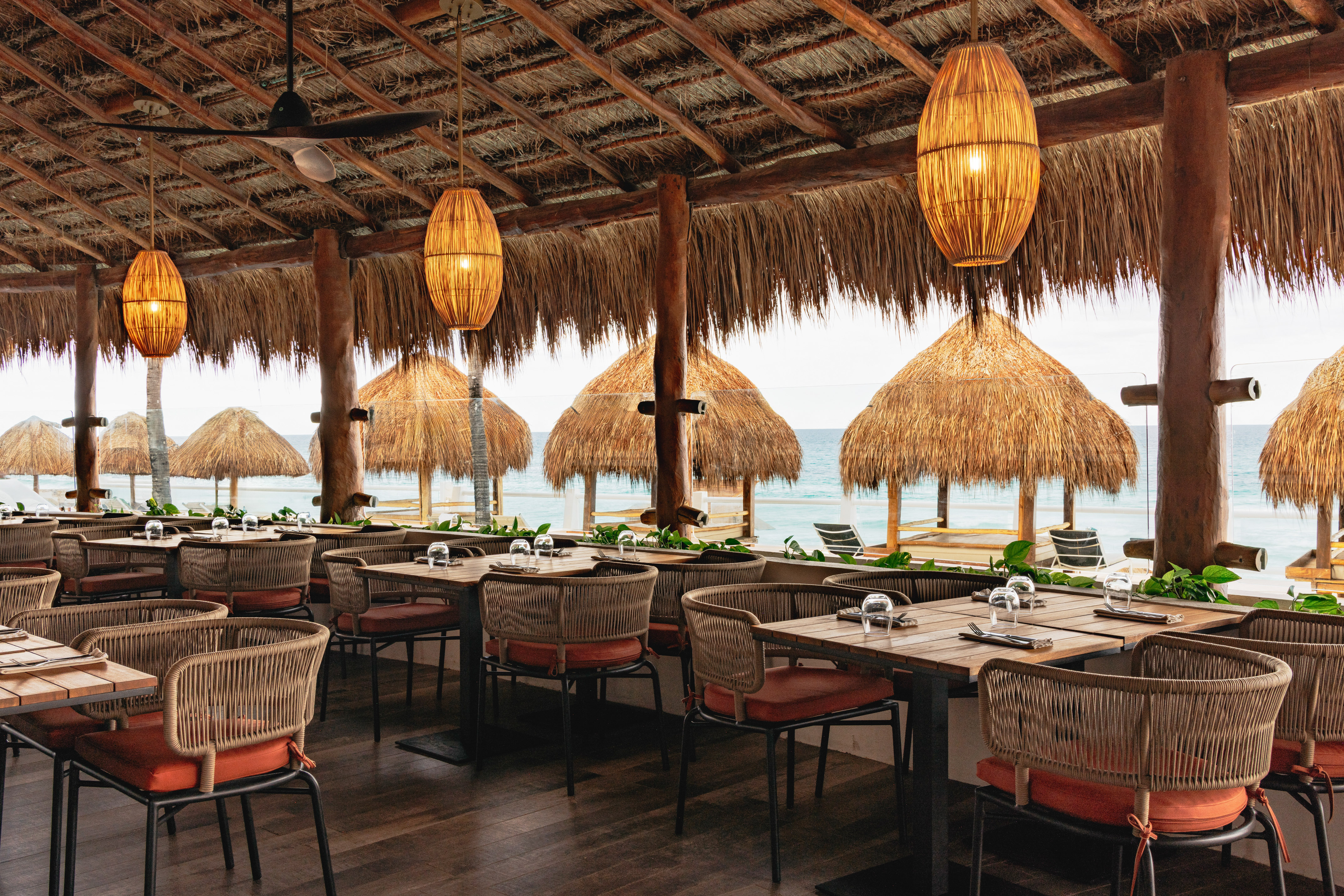 Beachfront palapa restaurant with ocean view, woven lamps, and set dining tables.