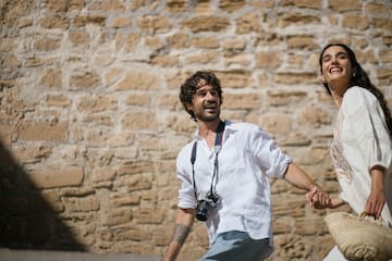 Happy couple holding hands walking outdoors by stone wall. Man has camera.