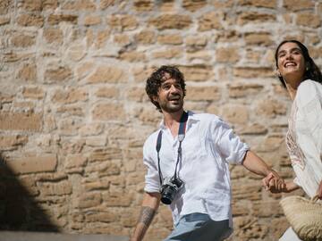 Happy couple holding hands walking outdoors by stone wall. Man has camera.