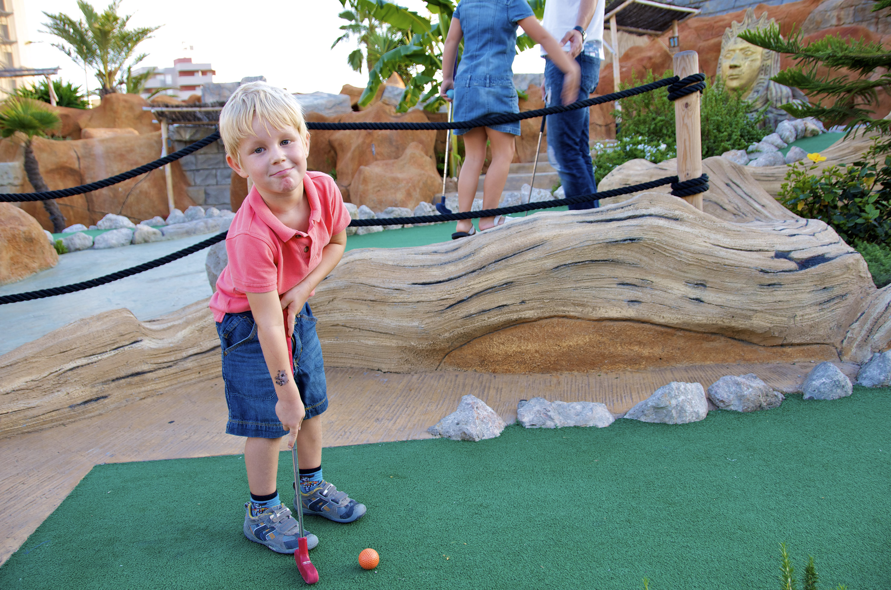 Smiling blond boy plays mini golf outdoors with a red putter on a green course.