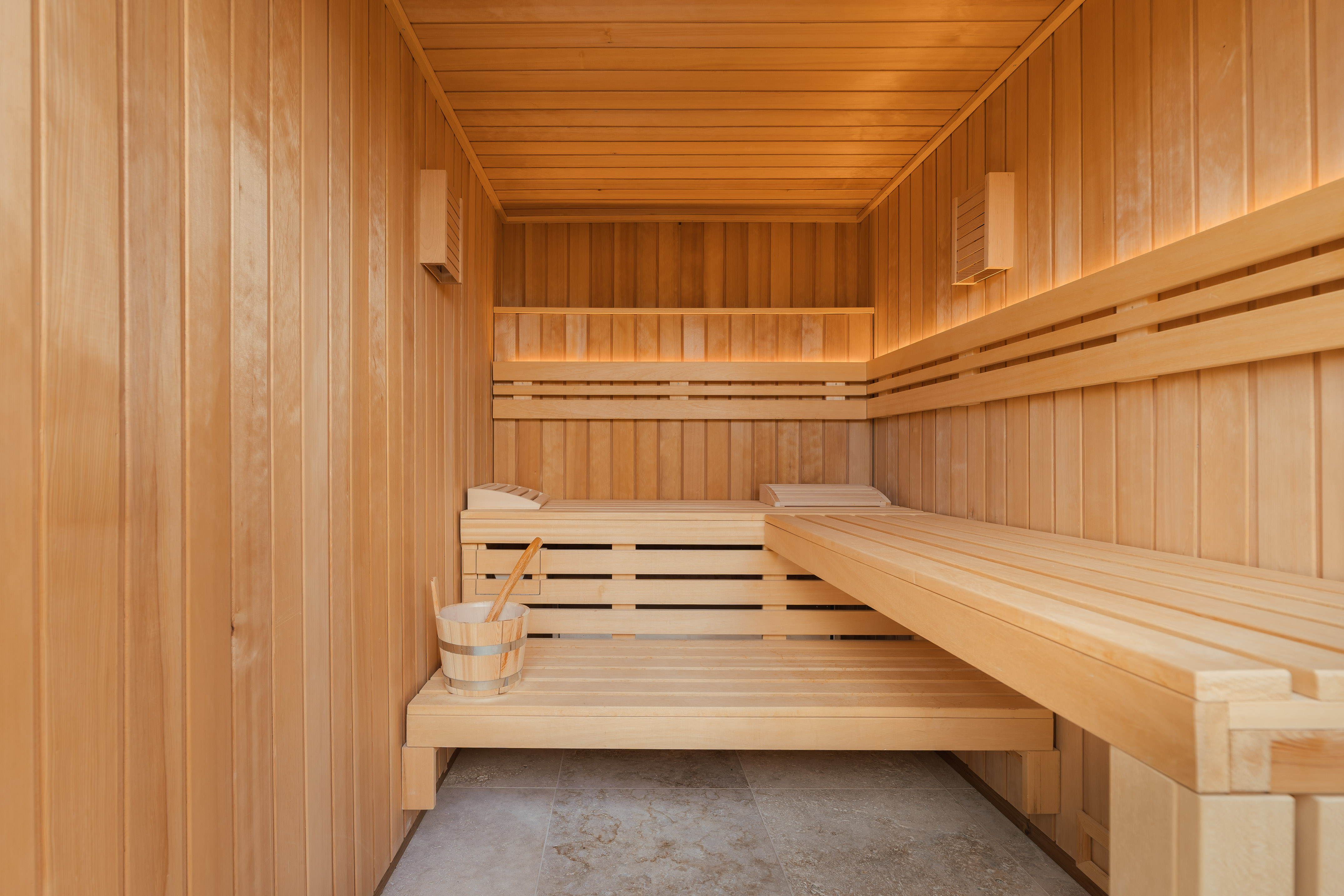 Light wood sauna room with benches, water bucket, and indirect lighting.