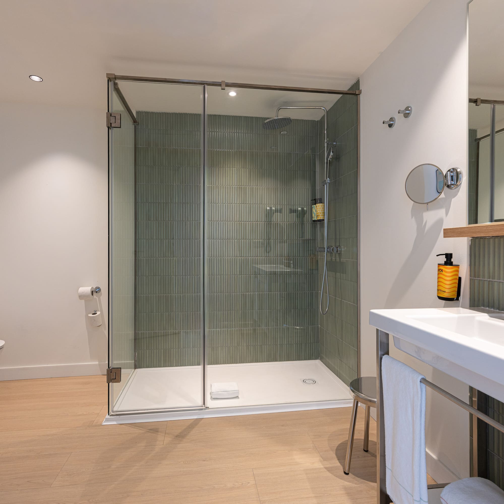 Modern hotel bathroom with glass shower, green tiled wall, toilet, and vanity.