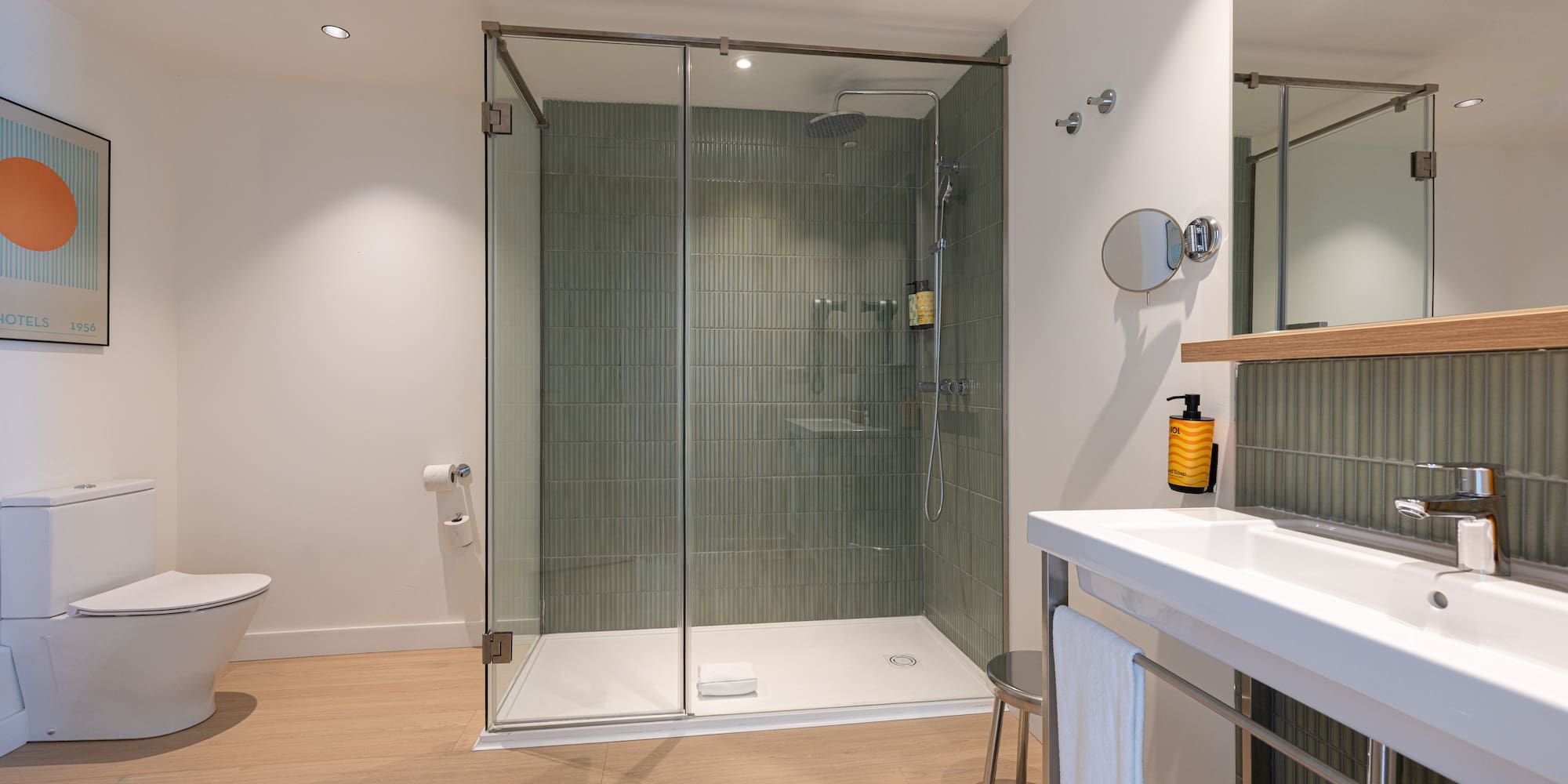 Modern hotel bathroom with glass shower, green tiled wall, toilet, and vanity.