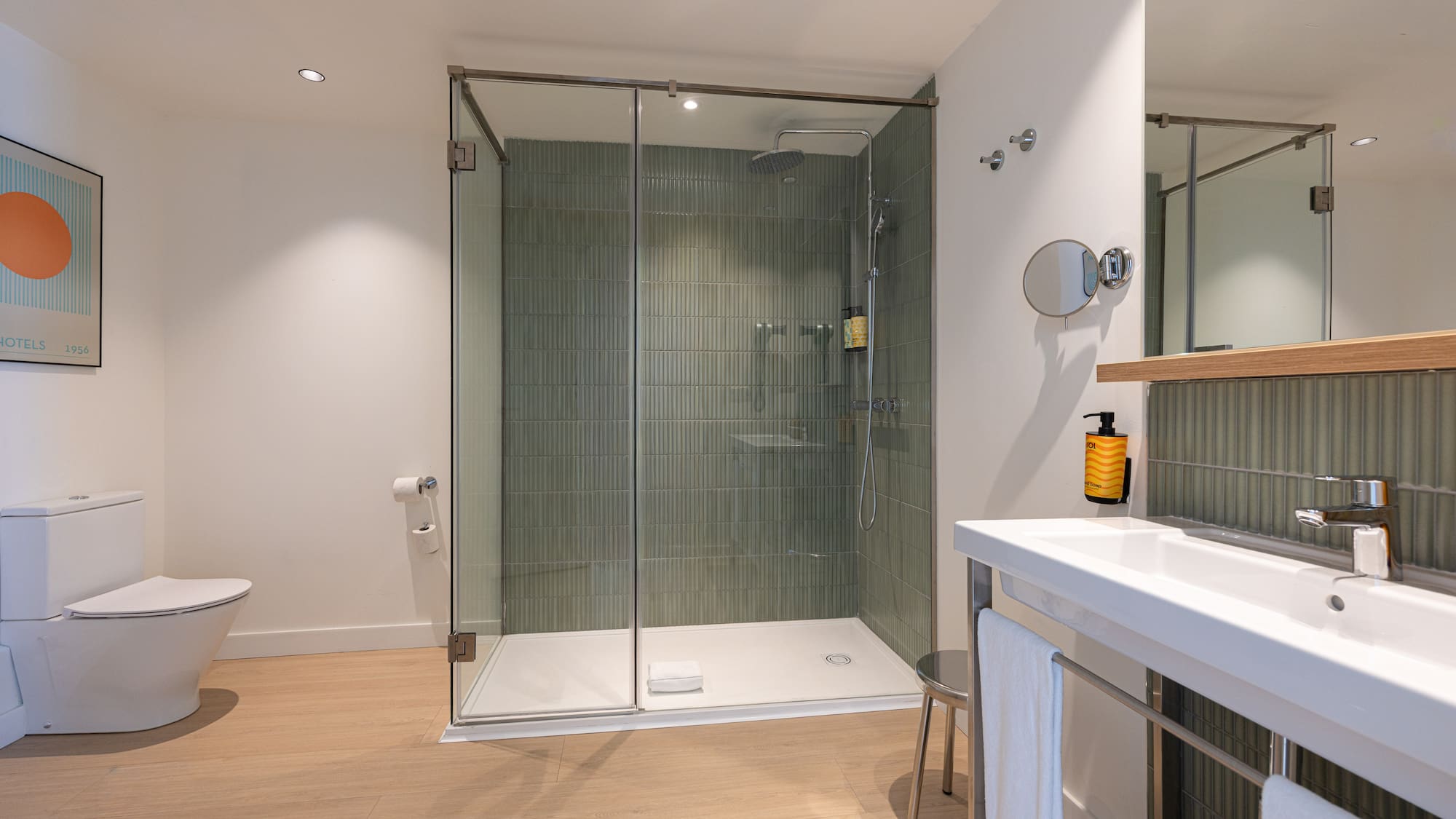 Modern hotel bathroom with glass shower, green tiled wall, toilet, and vanity.