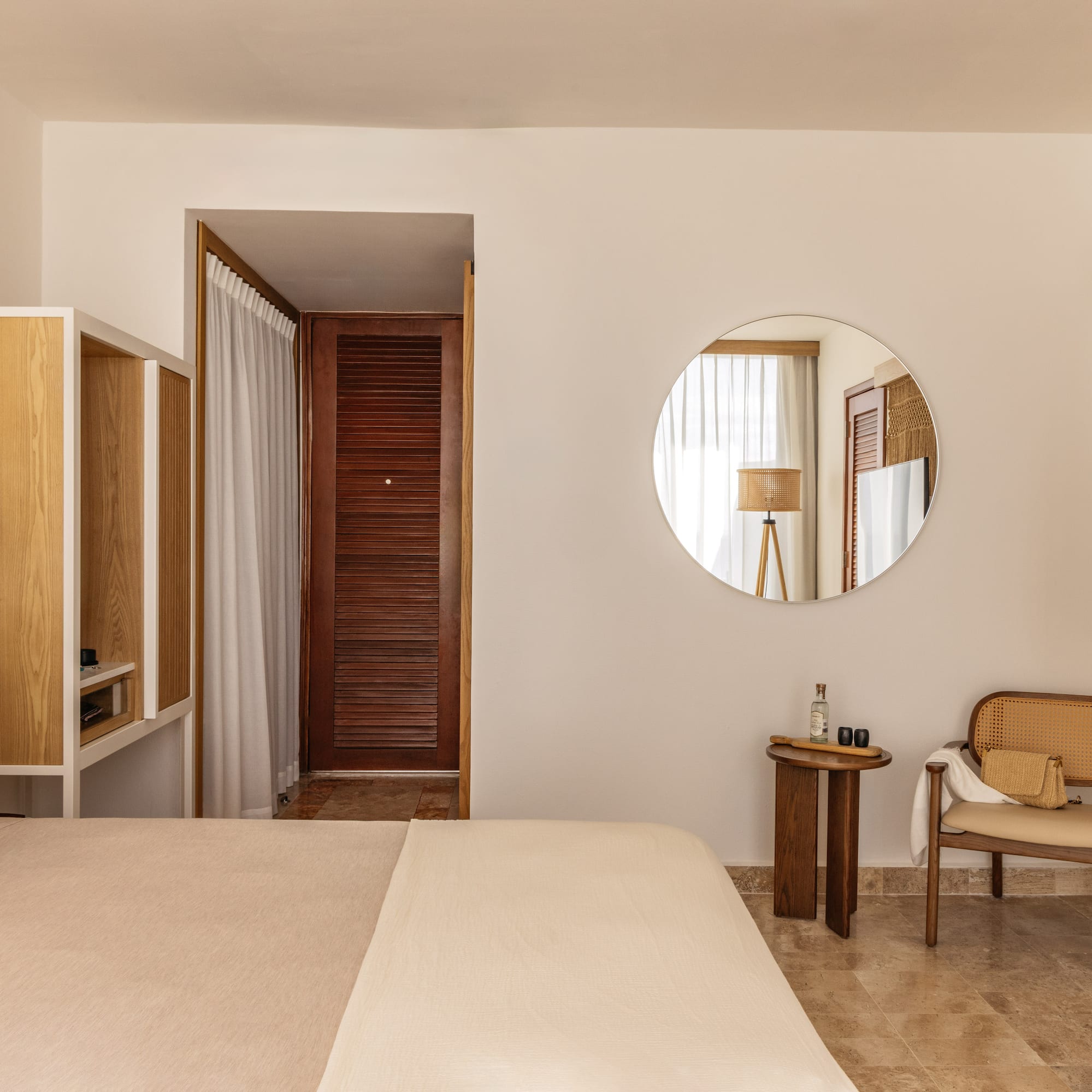 Cozy neutral hotel room with bed, wooden furniture, rattan chair, and round mirror.