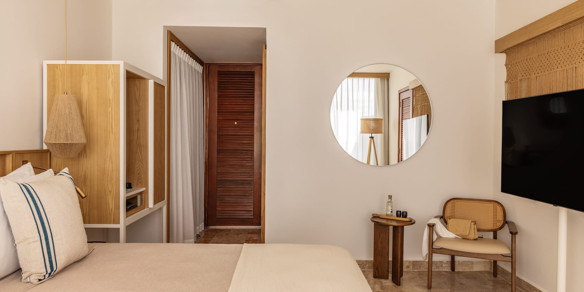 Cozy neutral hotel room with bed, wooden furniture, rattan chair, and round mirror.