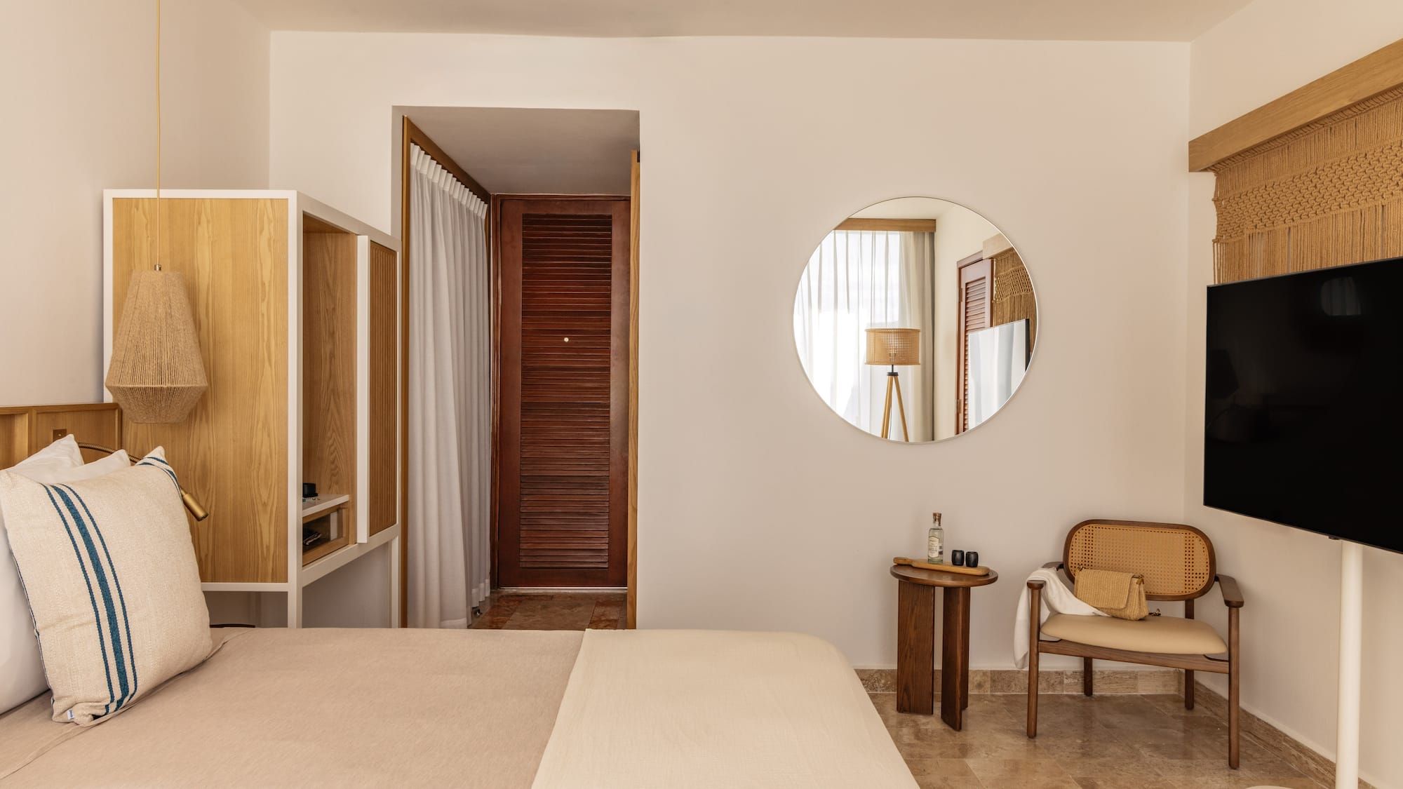 Cozy neutral hotel room with bed, wooden furniture, rattan chair, and round mirror.
