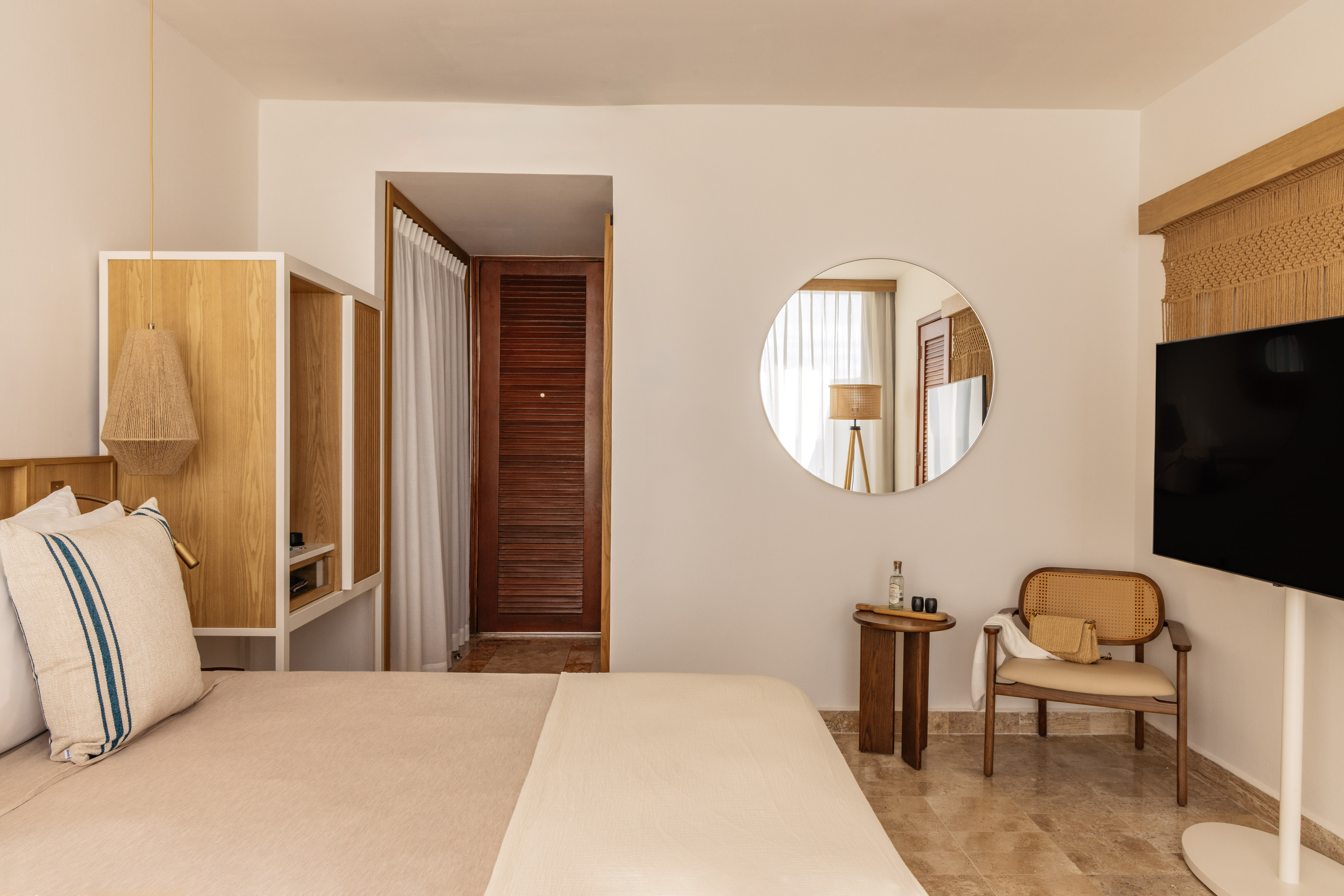 Cozy neutral hotel room with bed, wooden furniture, rattan chair, and round mirror.