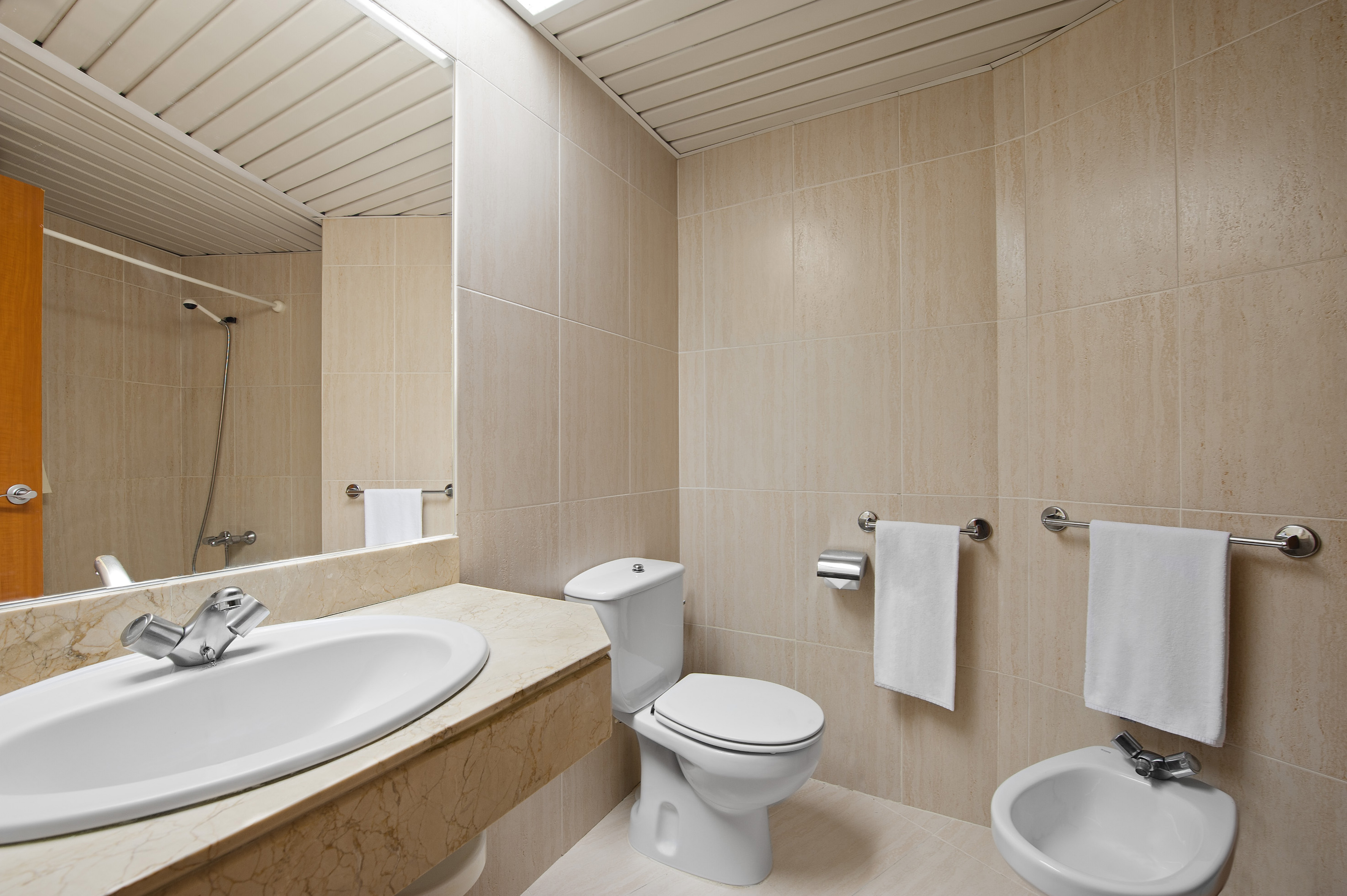 Clean beige tiled bathroom featuring sink, toilet, bidet, and mirror.