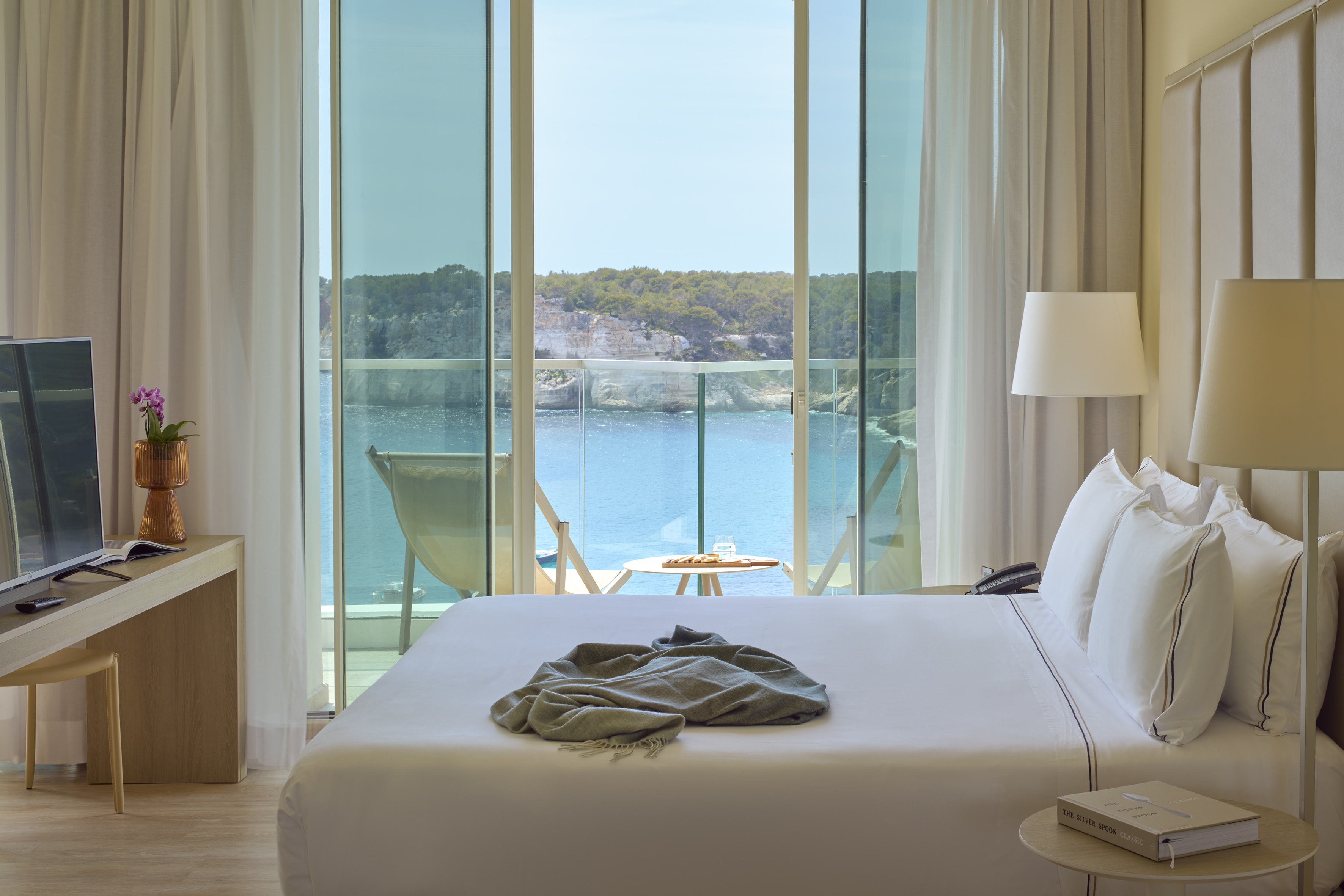 Modern hotel room with ocean view balcony, white bed, and rocky coast.