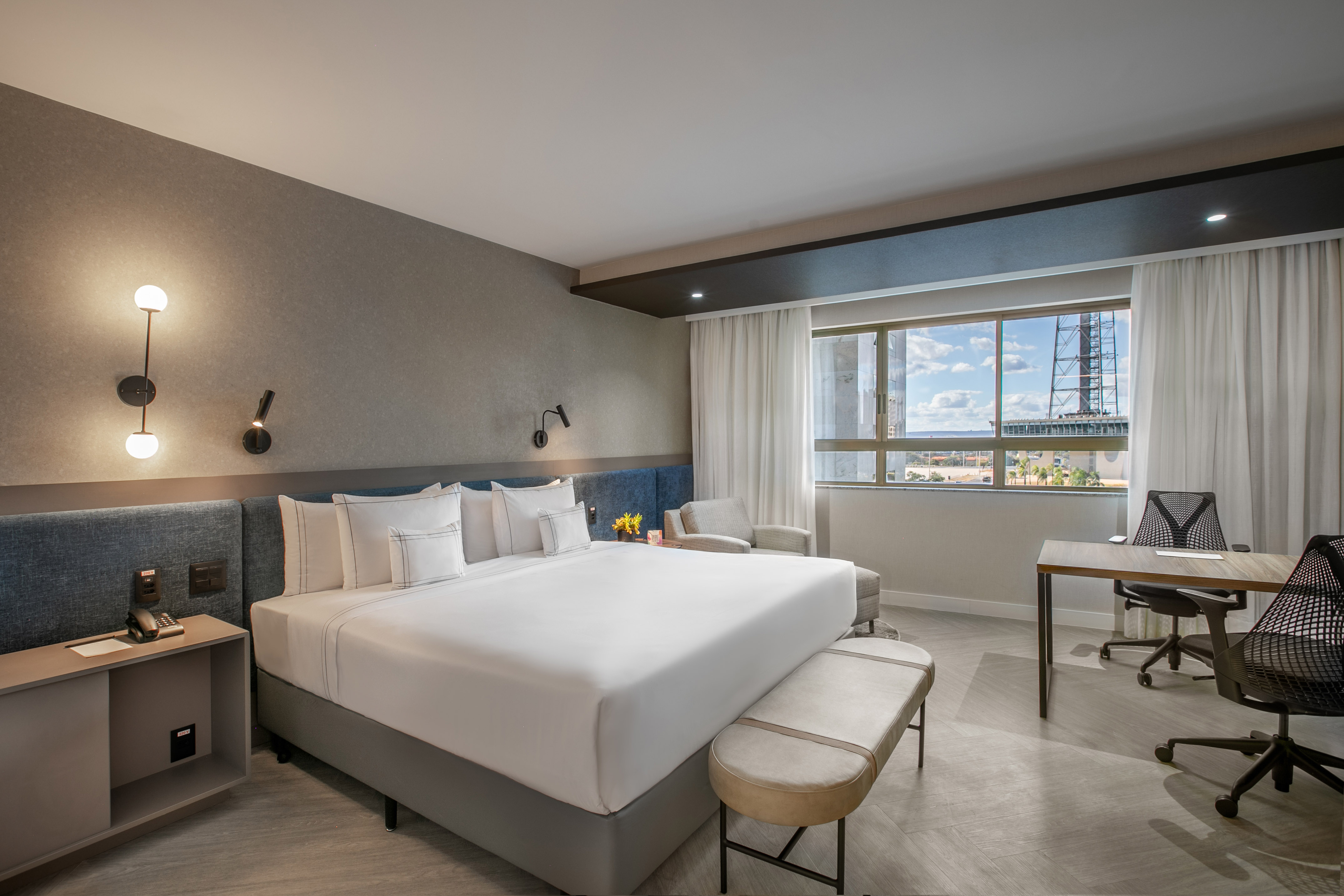 Modern hotel room with king bed, blue headboard, desk, and city view from window.