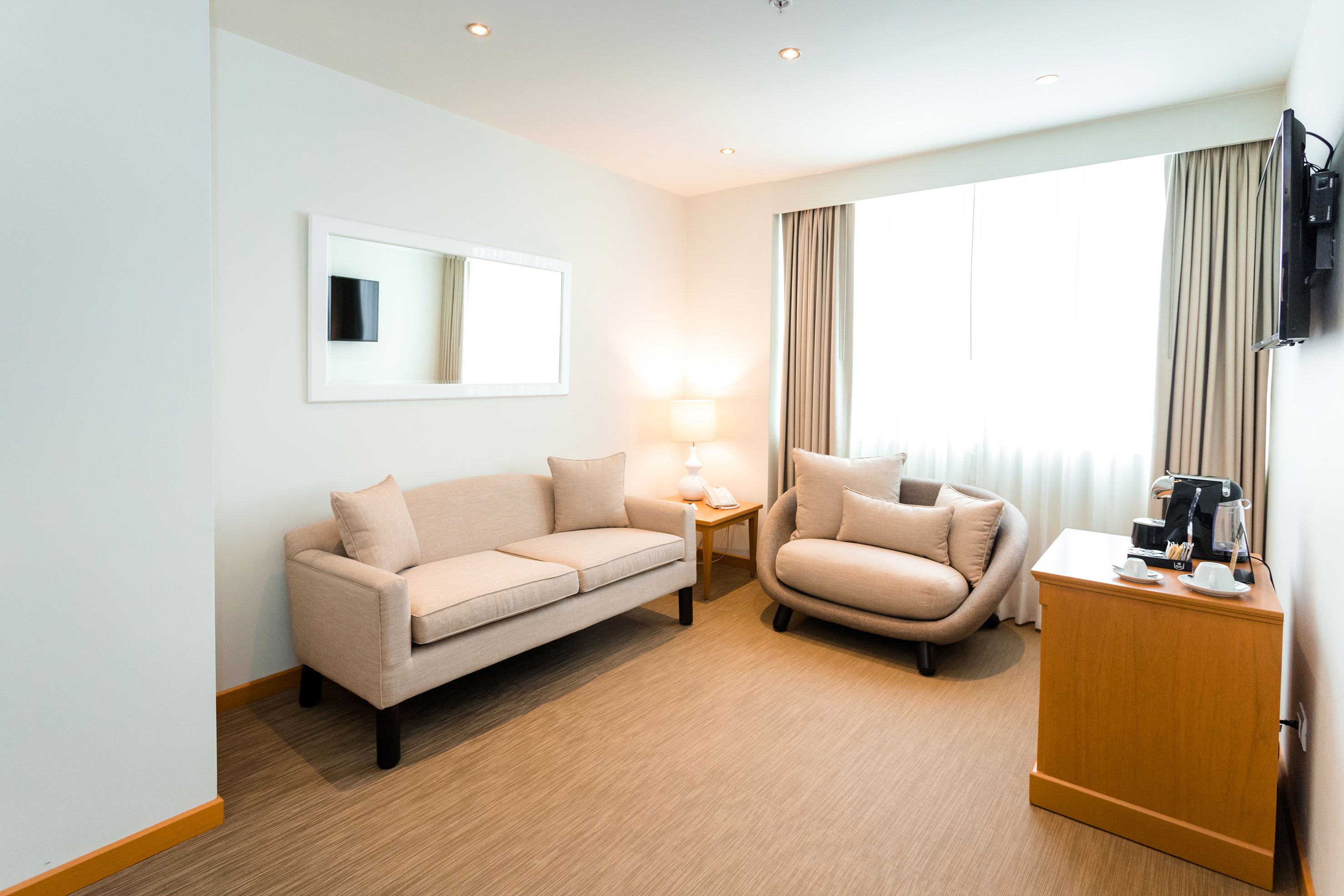 Comfortable hotel living area with beige sofa, armchair, coffee maker, and bright window.