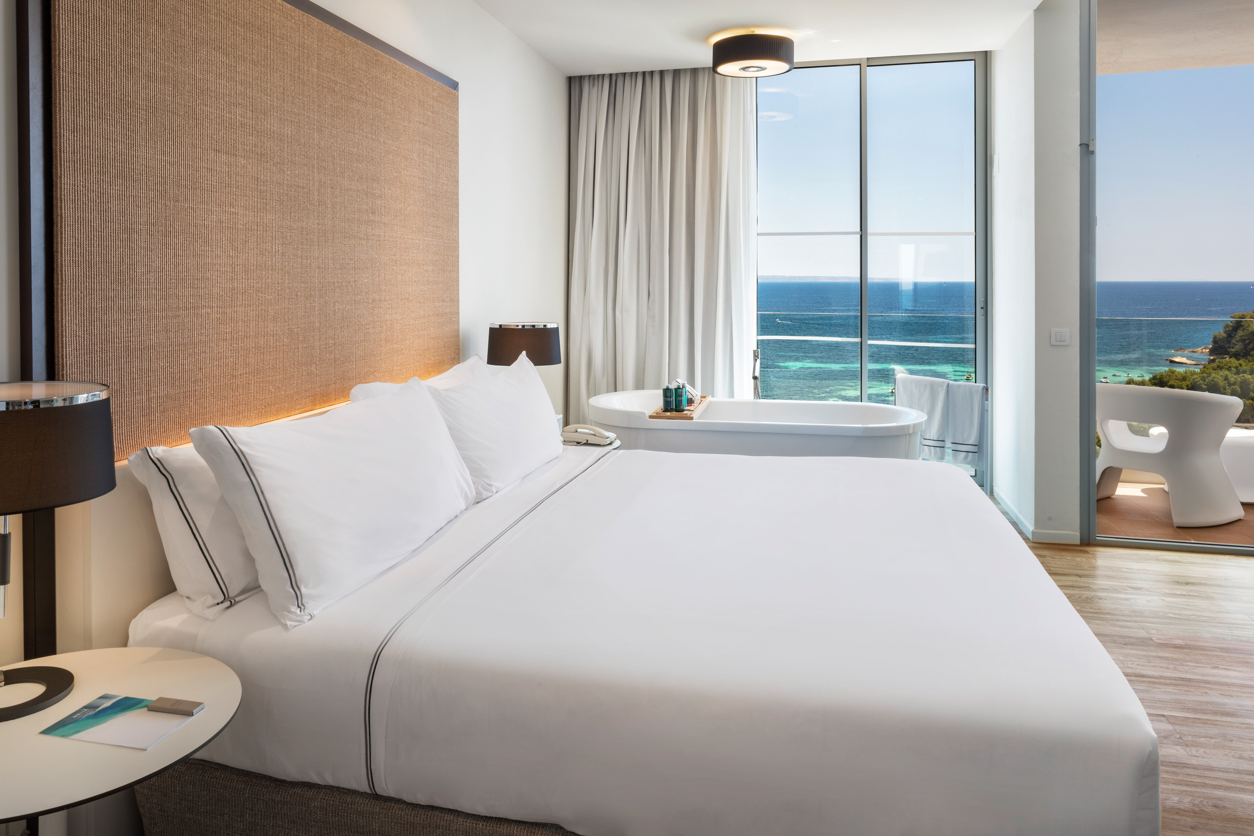 Modern luxury hotel room with ocean view, white bed, and freestanding bathtub.