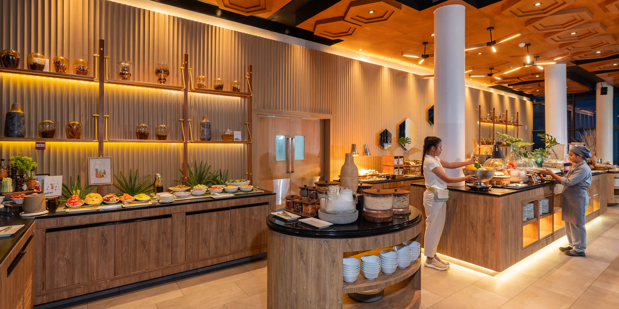 Modern hotel restaurant buffet with staff and varied food, warm wood interior.
