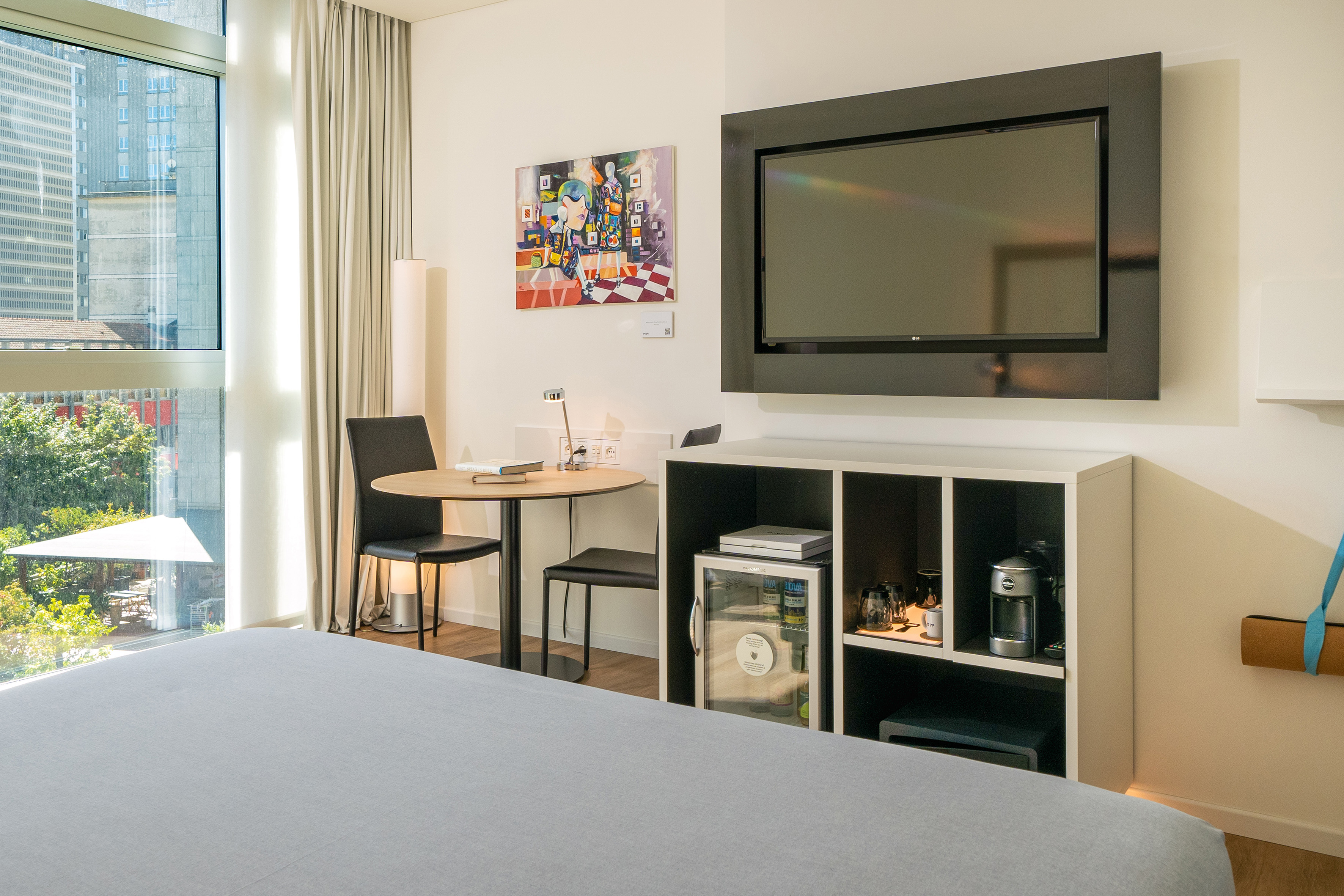 Modern hotel room with city view, bed, flat-screen TV, table, and mini-bar.