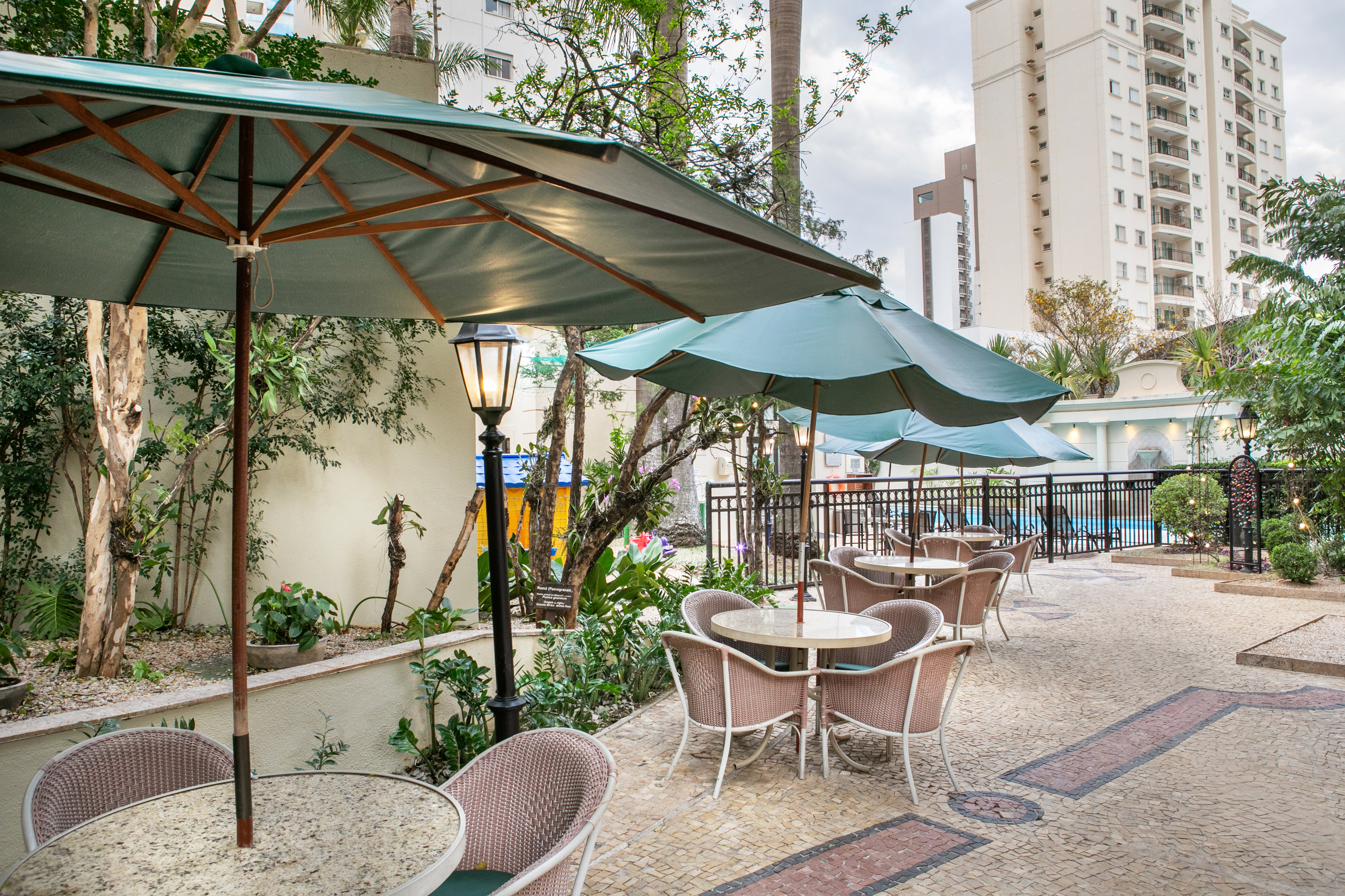 Outdoor cafe patio with green umbrellas, wicker furniture, lush garden, and pool.