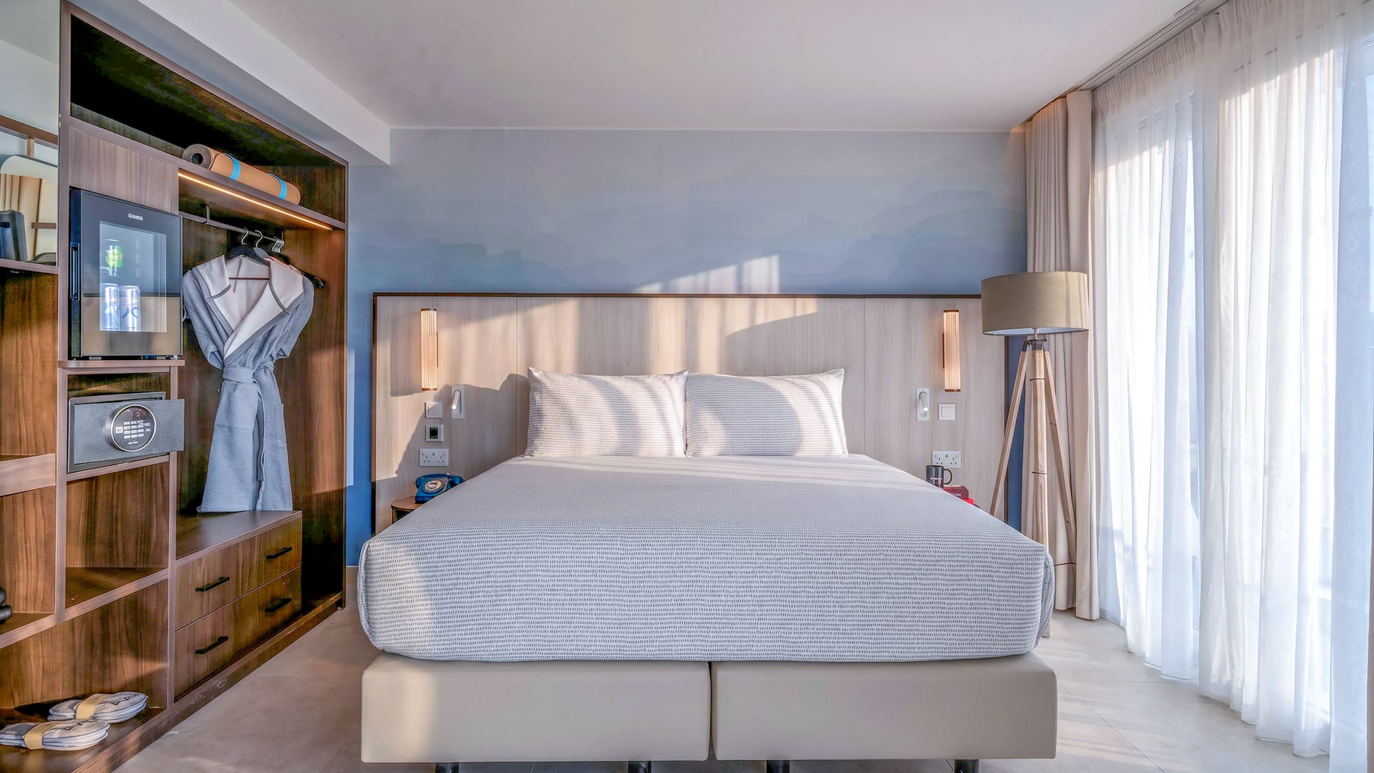 Bright, modern hotel bedroom with king bed, built-in storage, and sheer window curtains.