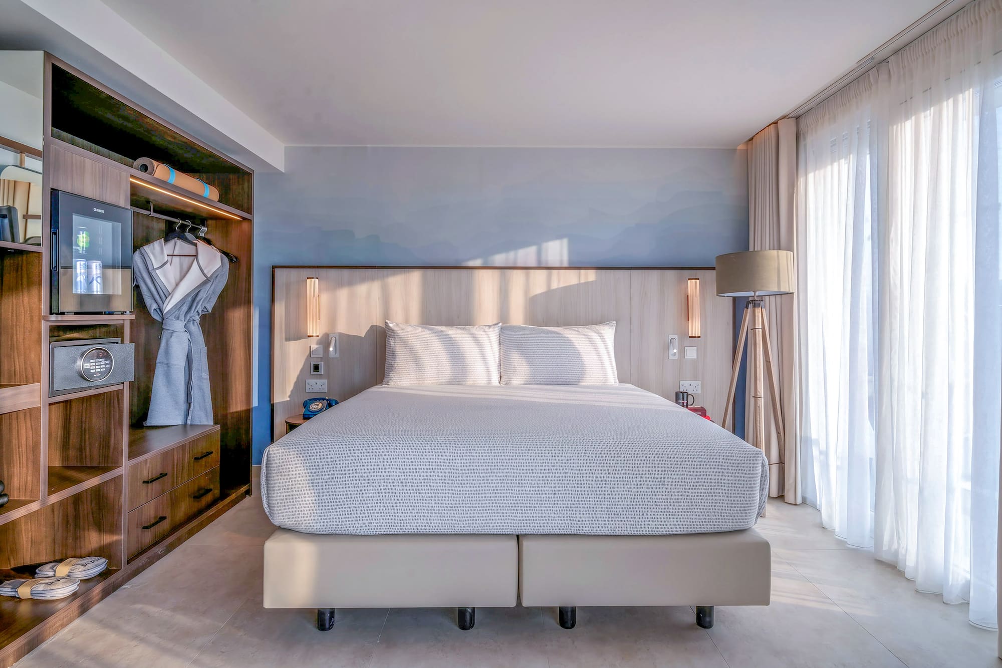 Bright, modern hotel bedroom with king bed, built-in storage, and sheer window curtains.