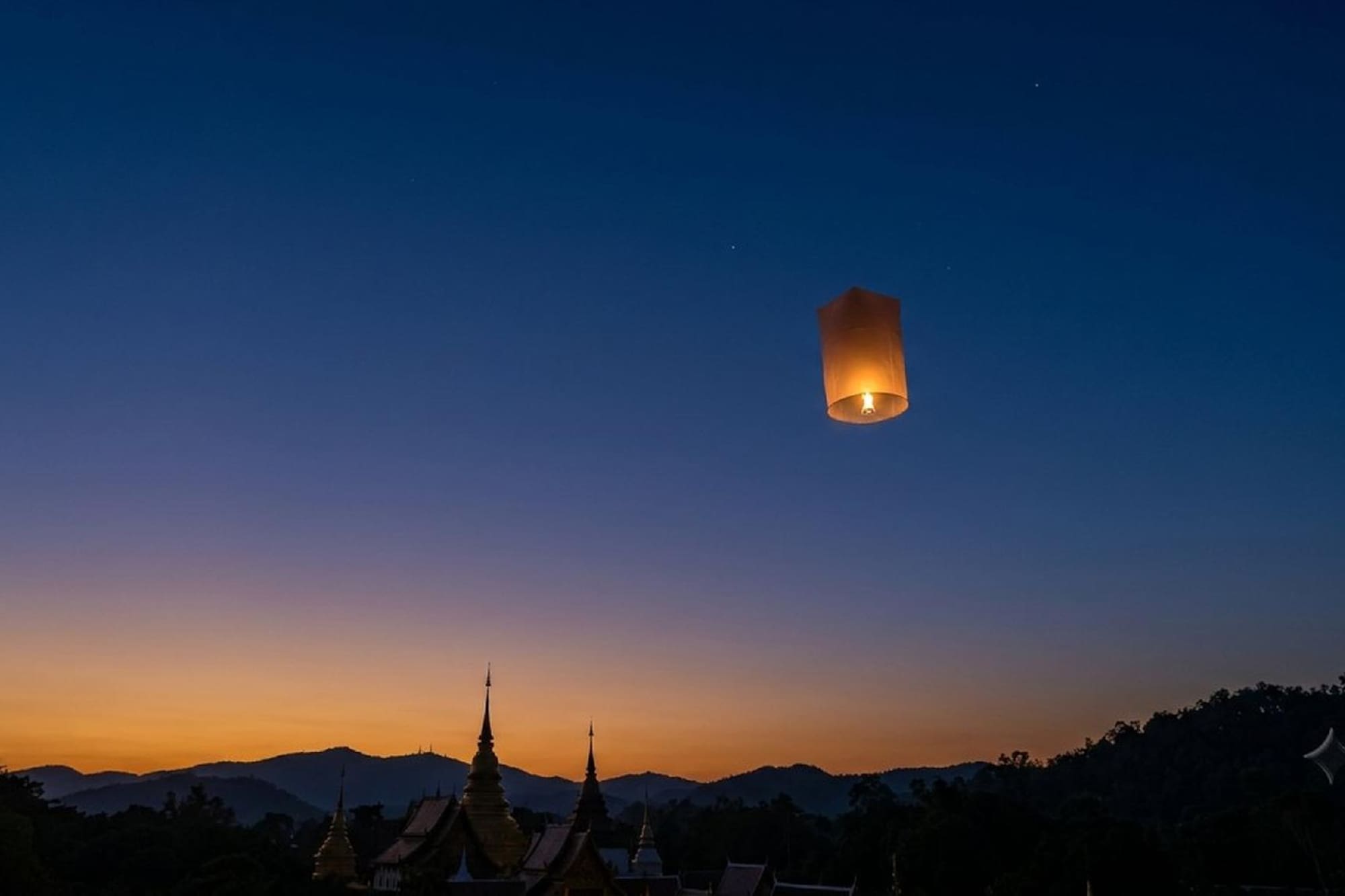 Glowing sky lantern above temple spires and mountains at sunset.