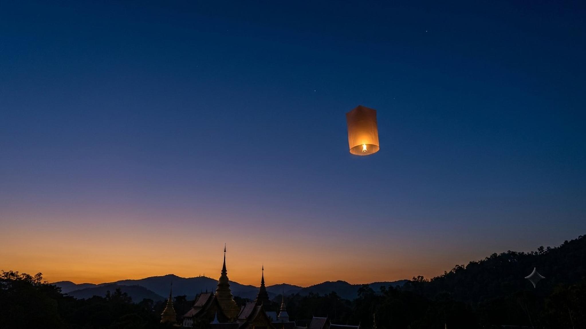 Glowing sky lantern above temple spires and mountains at sunset.