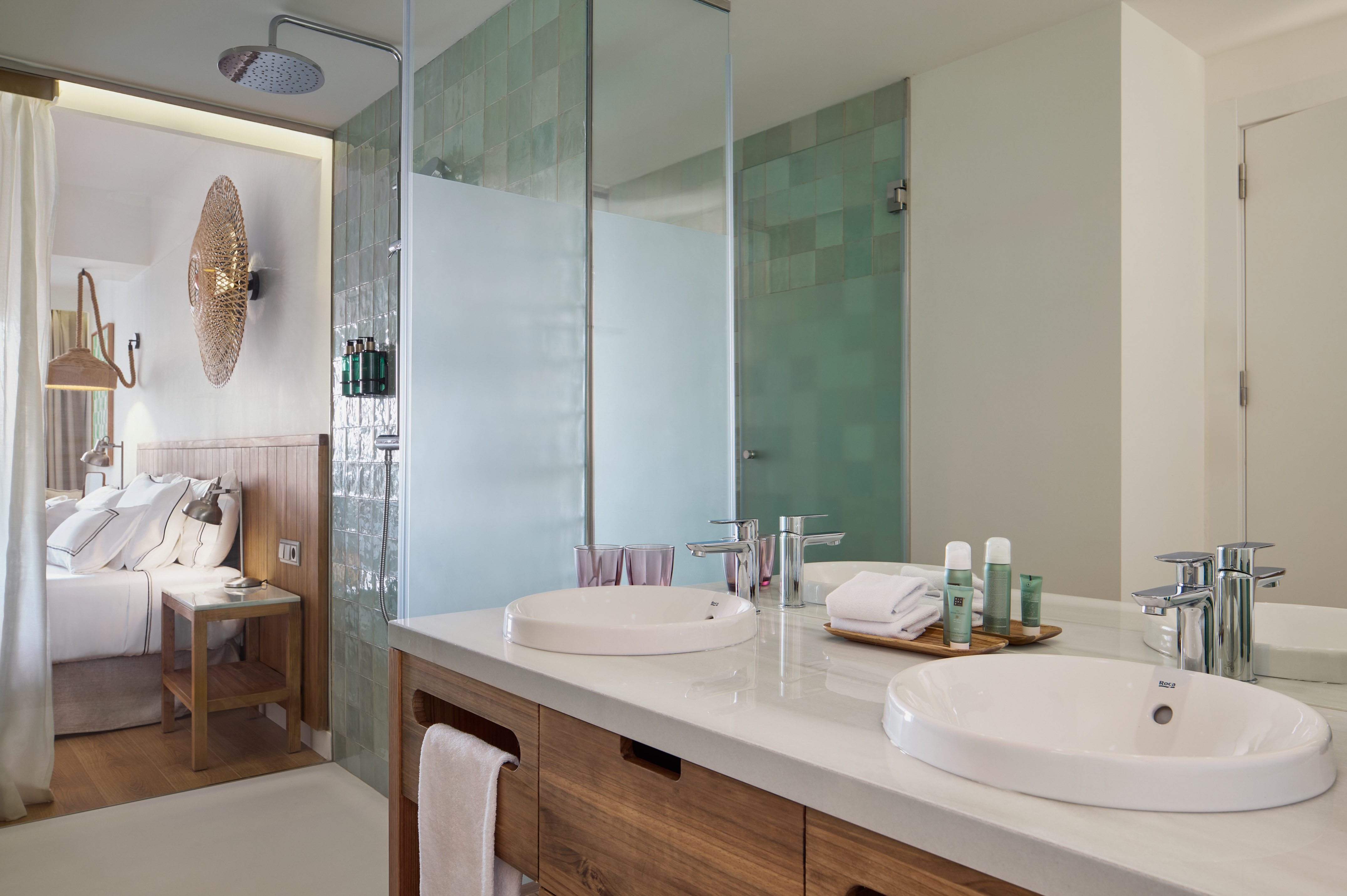 Modern hotel bathroom with double vanity, green tile shower, and bedroom view.