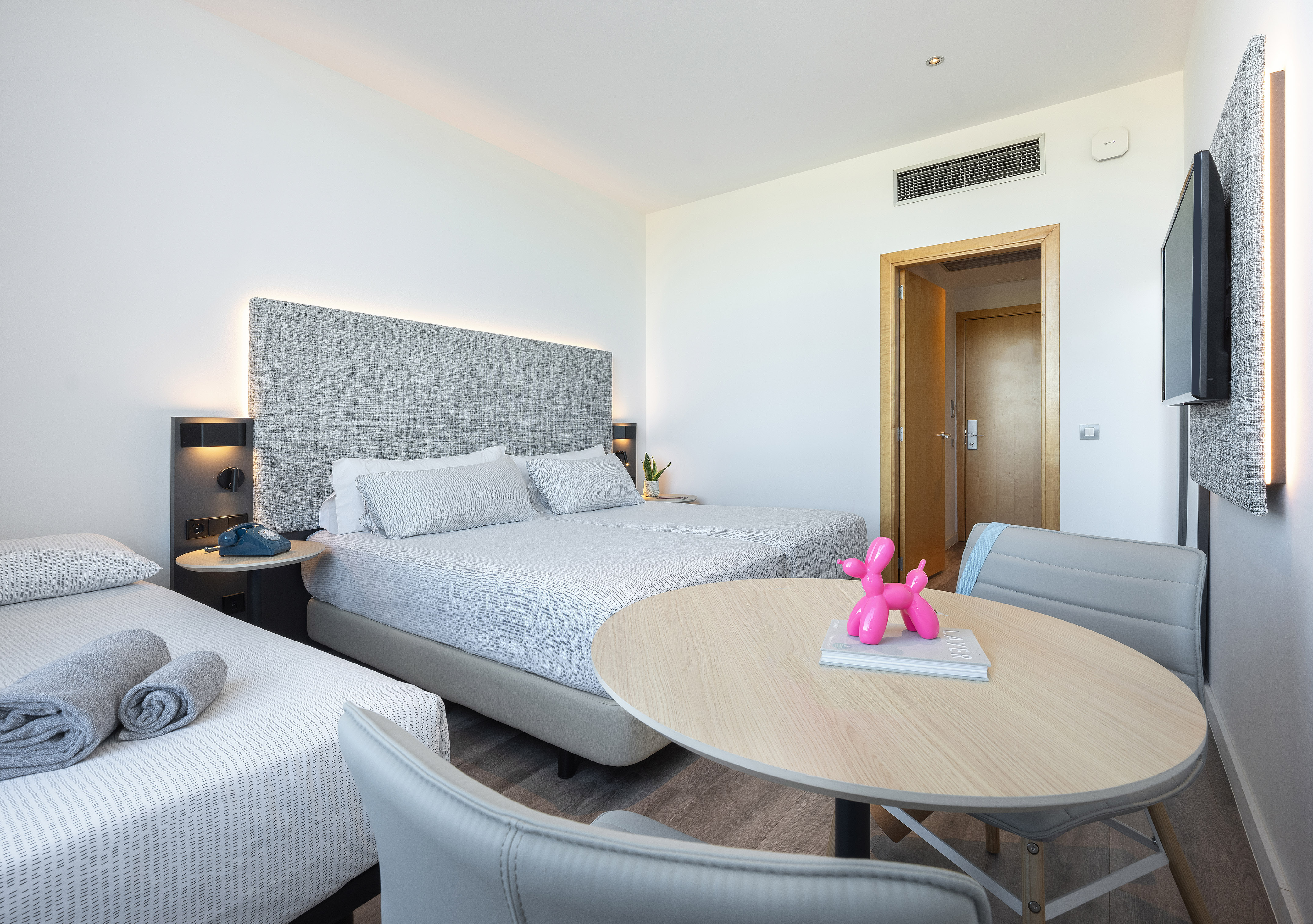 Bright modern hotel room with three beds, table, chairs, and pink balloon dog.