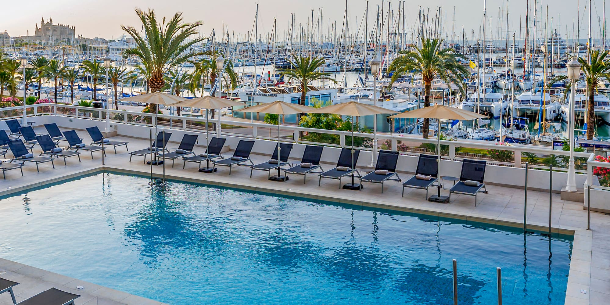Hotel pool, sun loungers, palm trees, and yacht marina with Palma Cathedral.