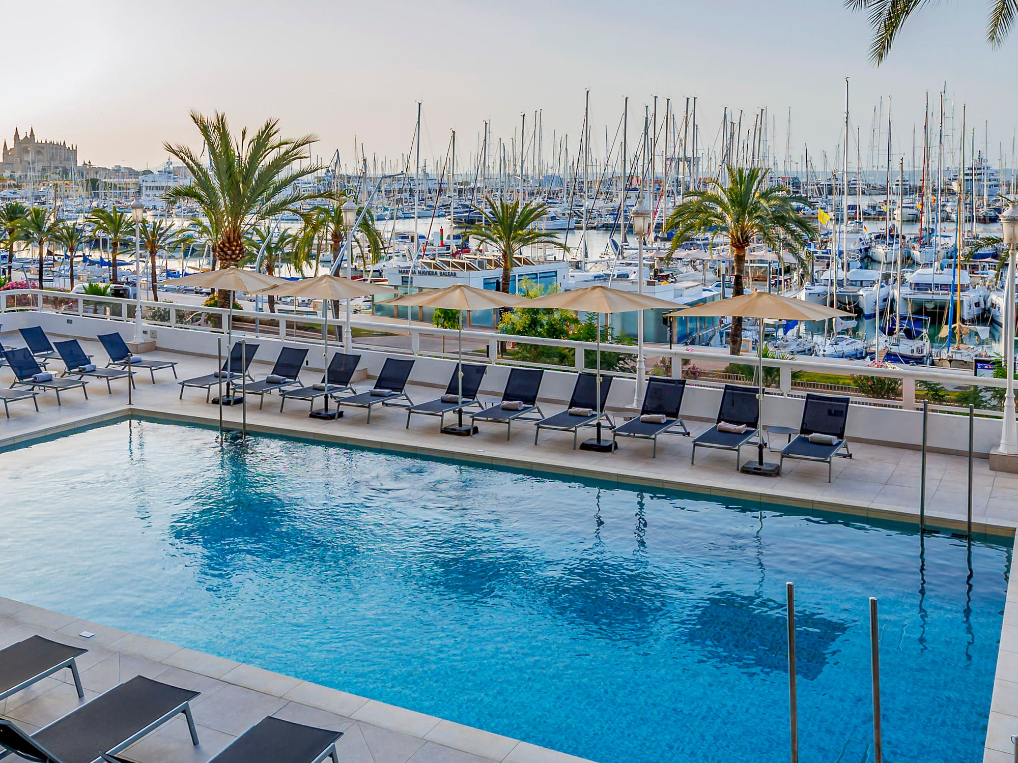 Piscina de hotel con tumbonas, vistas al puerto deportivo y Catedral de Palma.