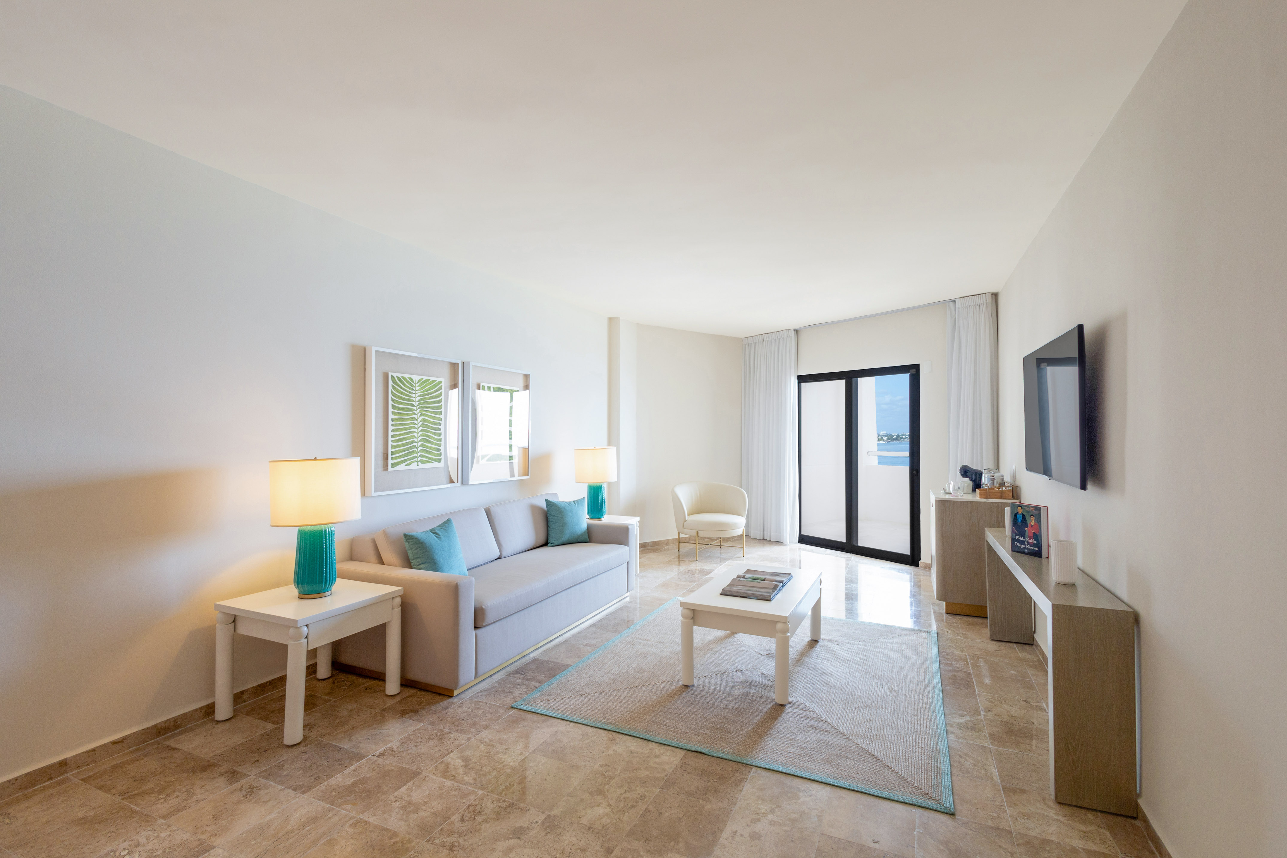 Bright hotel suite living room with sofa, TV, turquoise accents, and balcony view.