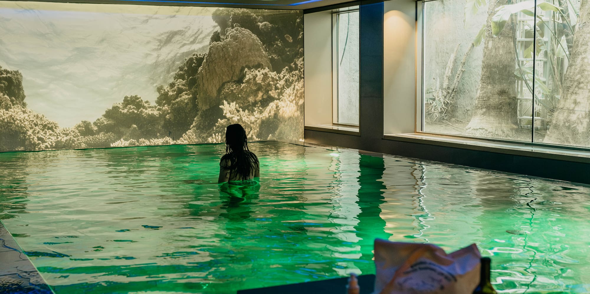 Woman in green lit indoor pool, coral reef mural, and tropical window view.