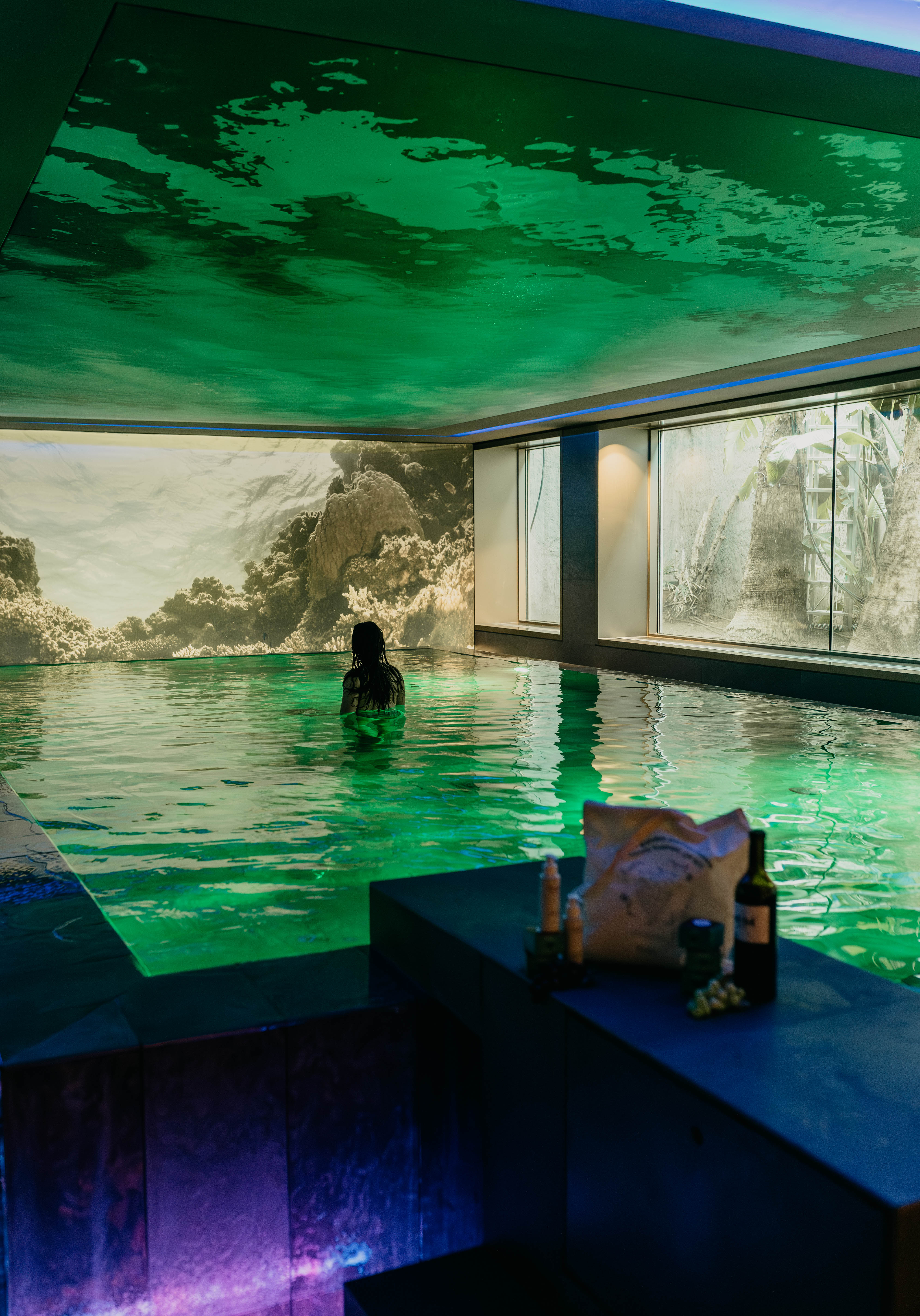 Woman in green lit indoor pool, coral reef mural, and tropical window view.