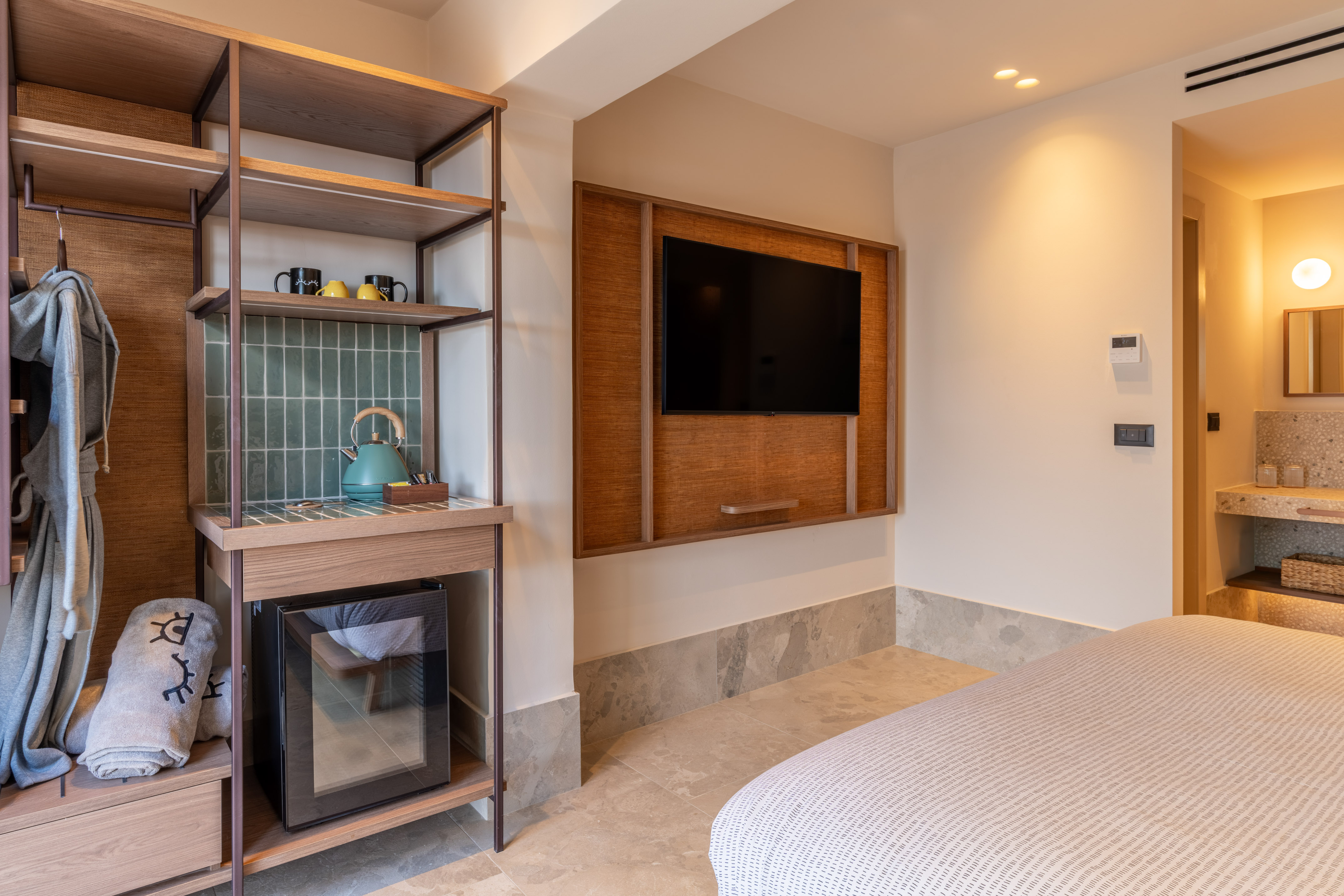 Modern hotel room with open wardrobe, minibar, kettle, and wall-mounted TV.