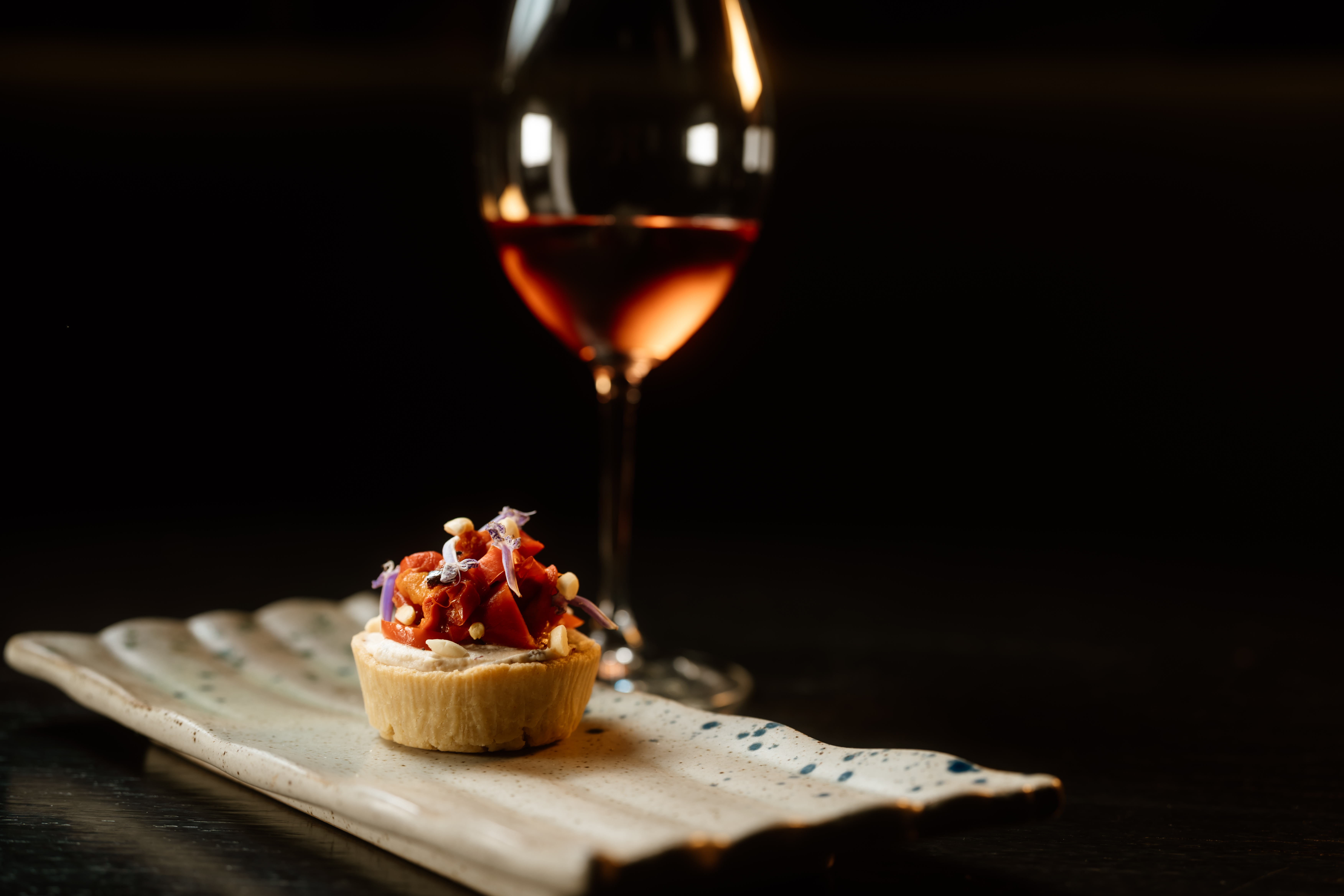 Gourmet tartlet with red filling, nuts, and flowers on plate; rosé wine glass.
