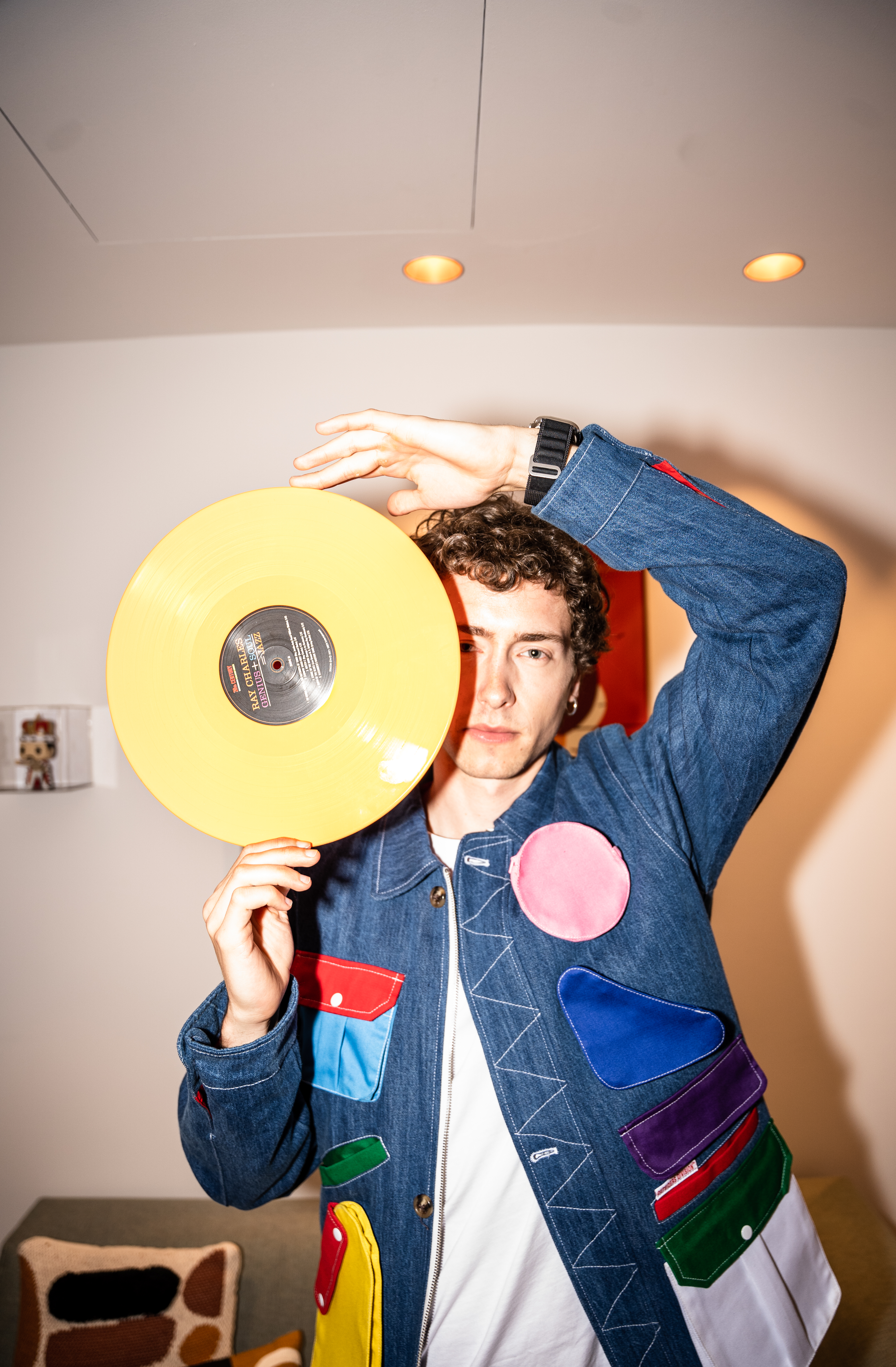 Man holding yellow Ray Charles vinyl record, wearing colorful patched denim jacket.