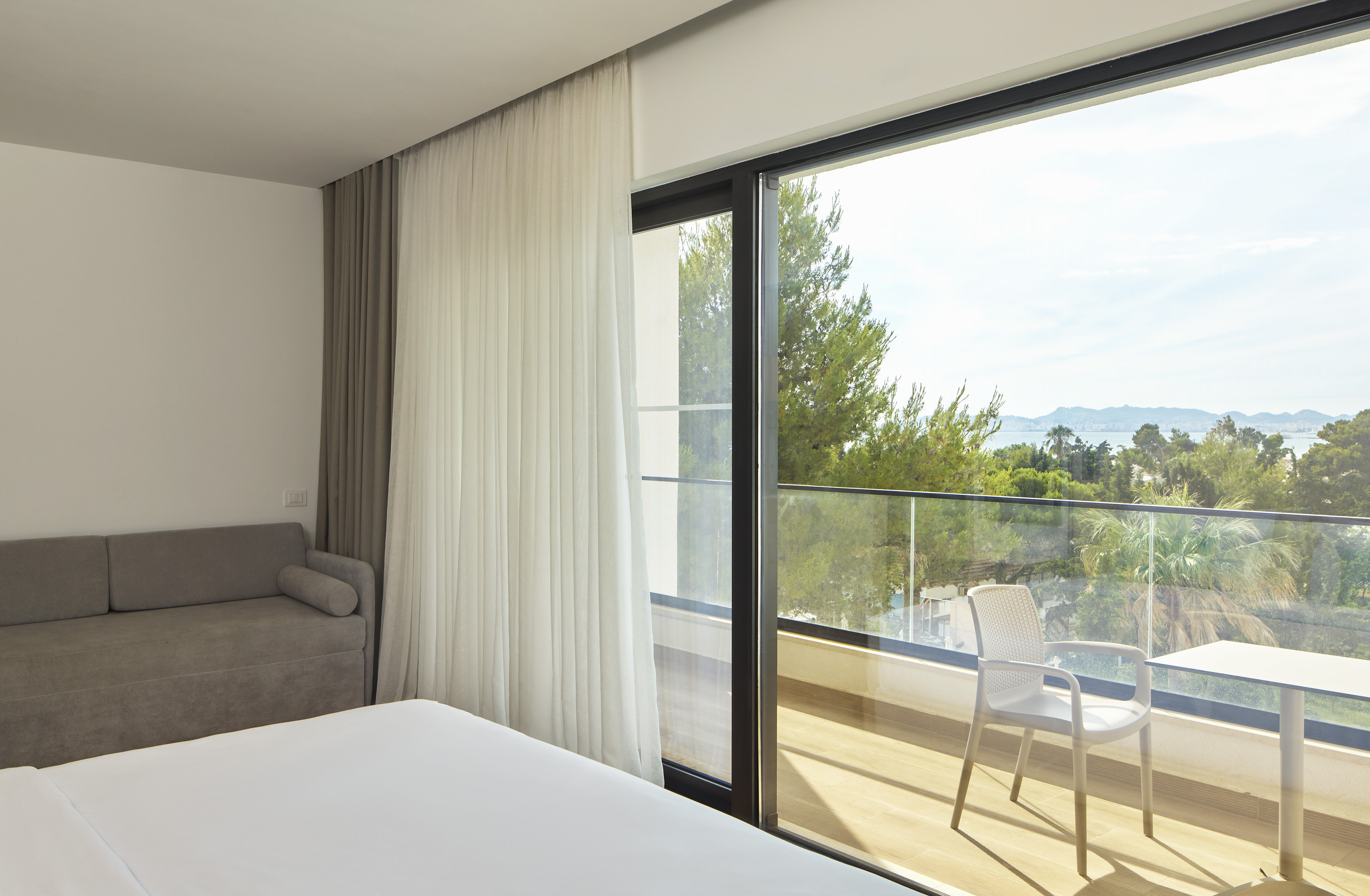 Bright hotel room with sea view balcony, bed, sofa, and outdoor seating.