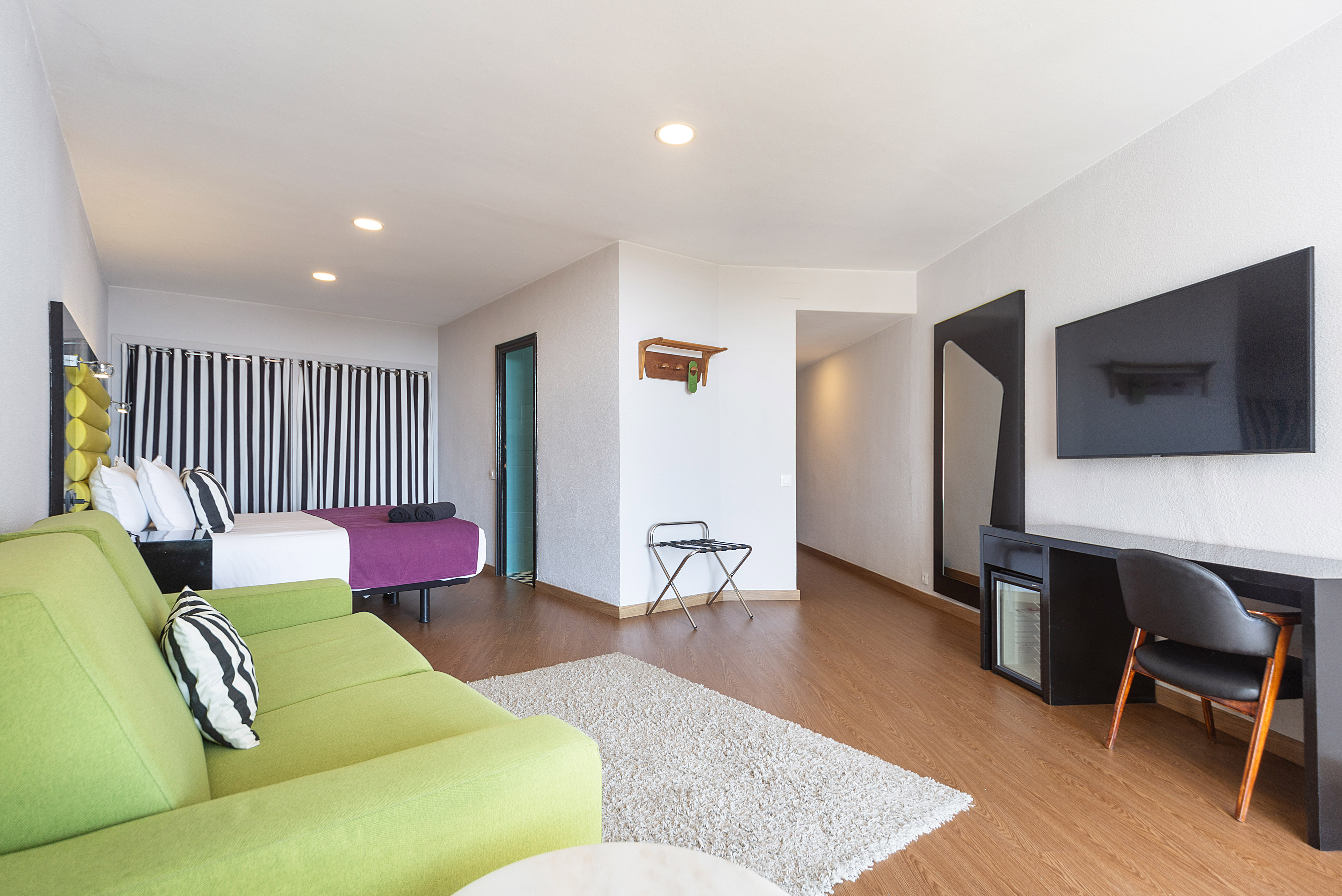 Stylish hotel suite with bed, bright green couch, desk, TV, and wood floor.