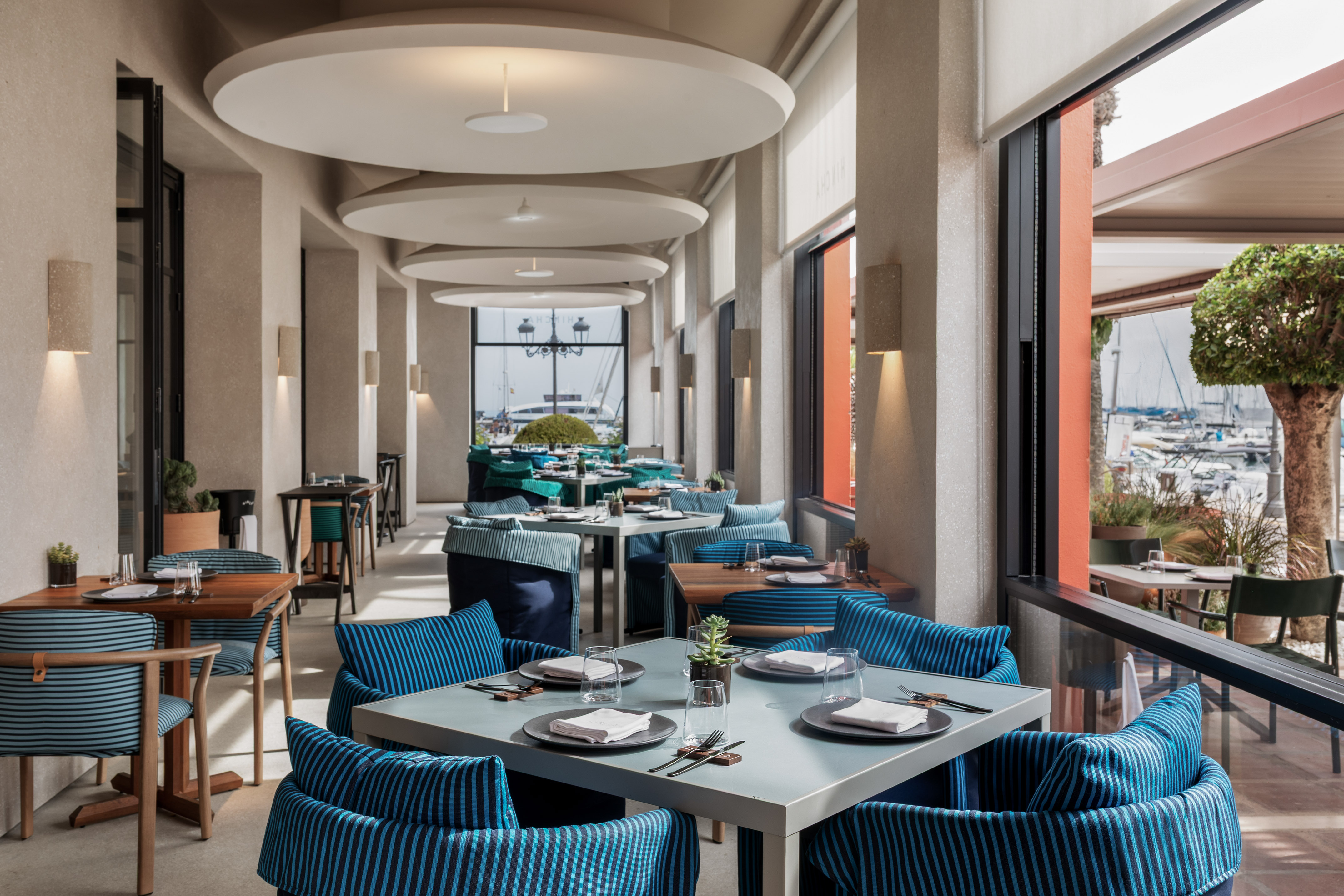 Modern marina restaurant, blue striped seating, set tables, waterfront view.