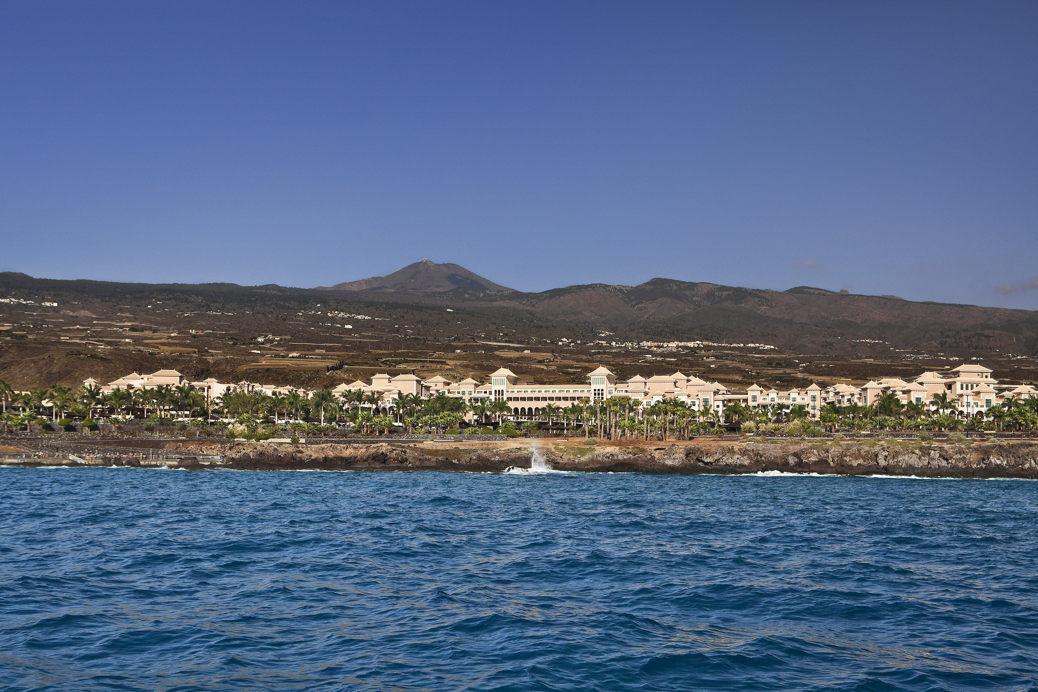 Tenerife resort on rocky coast, blue ocean, with Teide volcano in background.