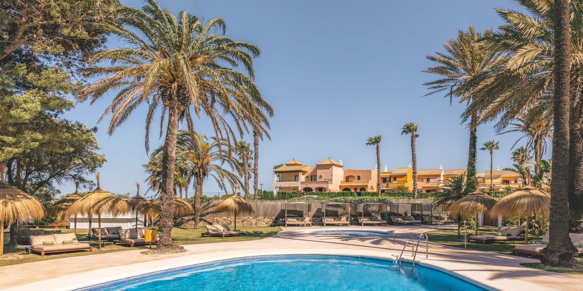Resort pool with palm trees, straw umbrellas, sun loungers, and hotel.