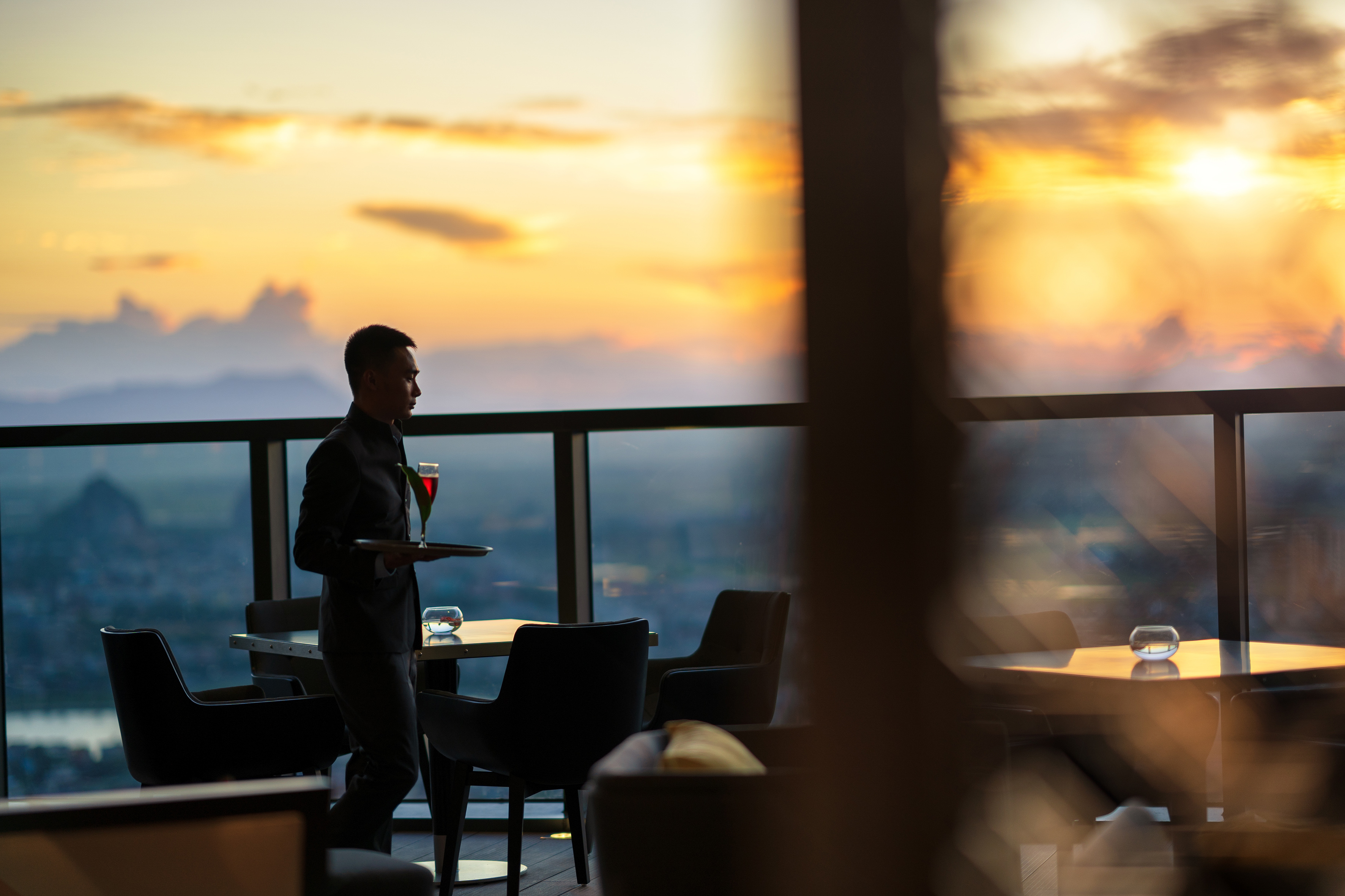 Rooftop bar server with cocktail, city sunset view.