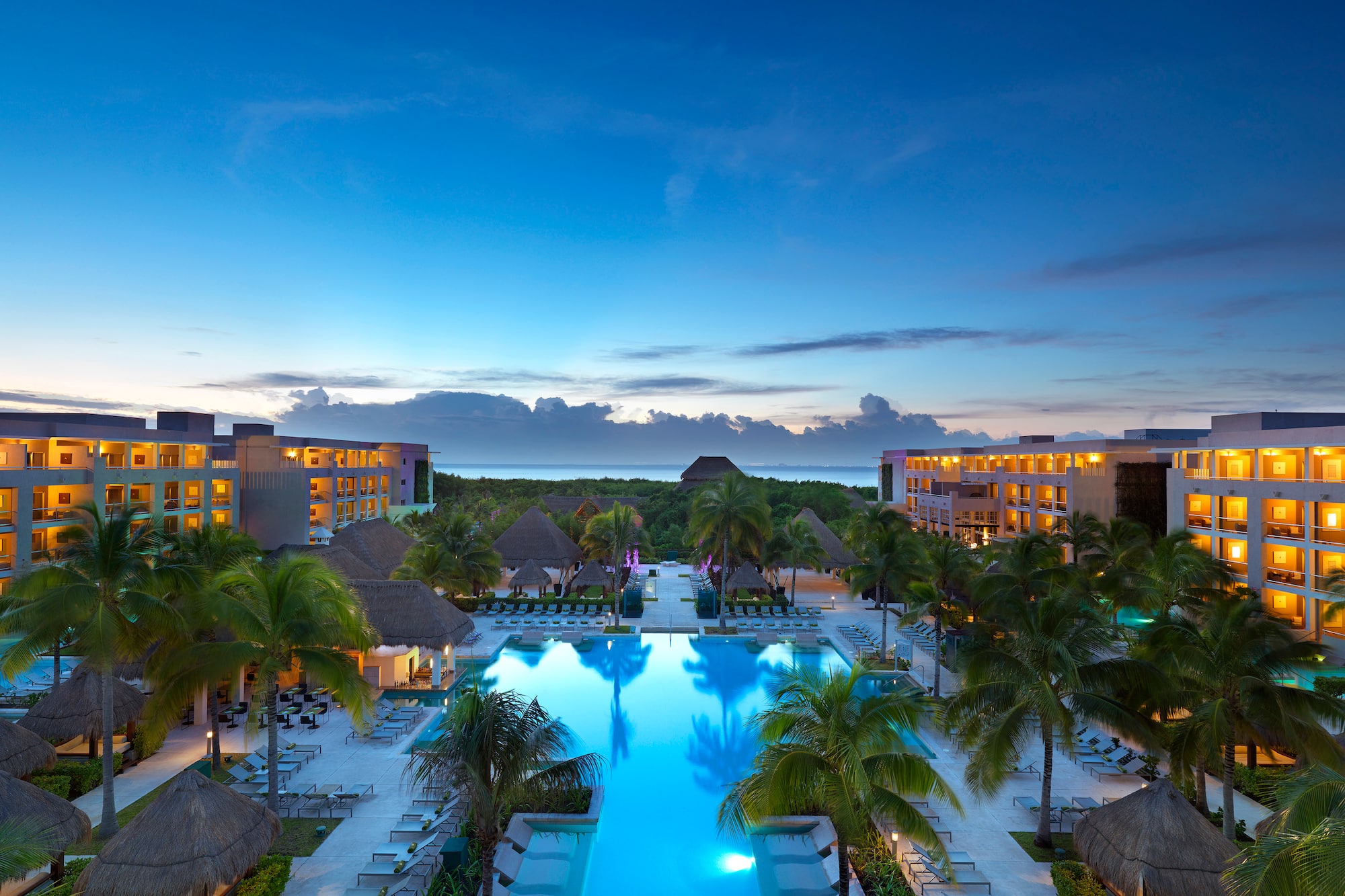 Tropical luxury resort, illuminated pool, palm trees, buildings at dusk.