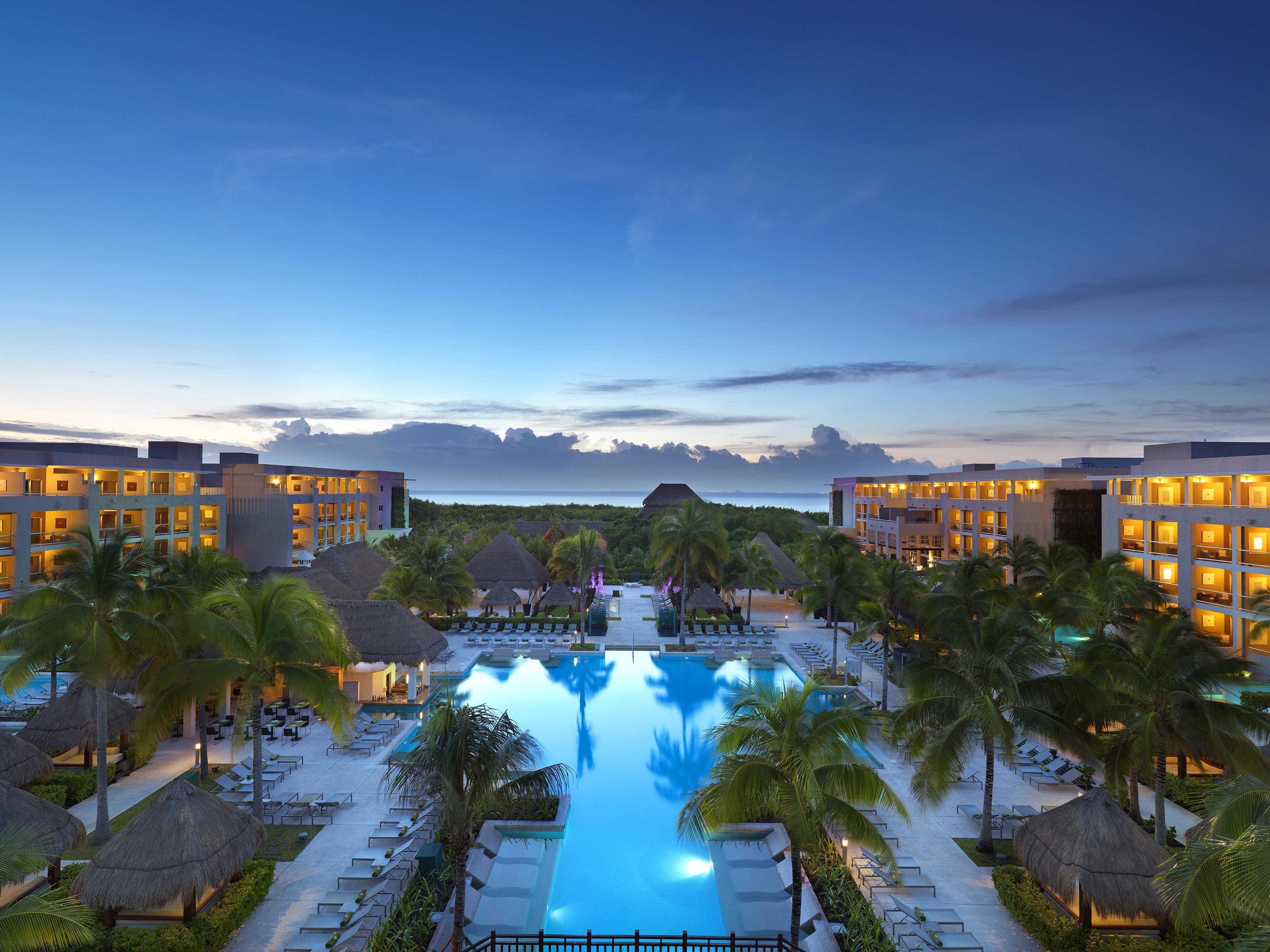 Tropical luxury resort, illuminated pool, palm trees, buildings at dusk.