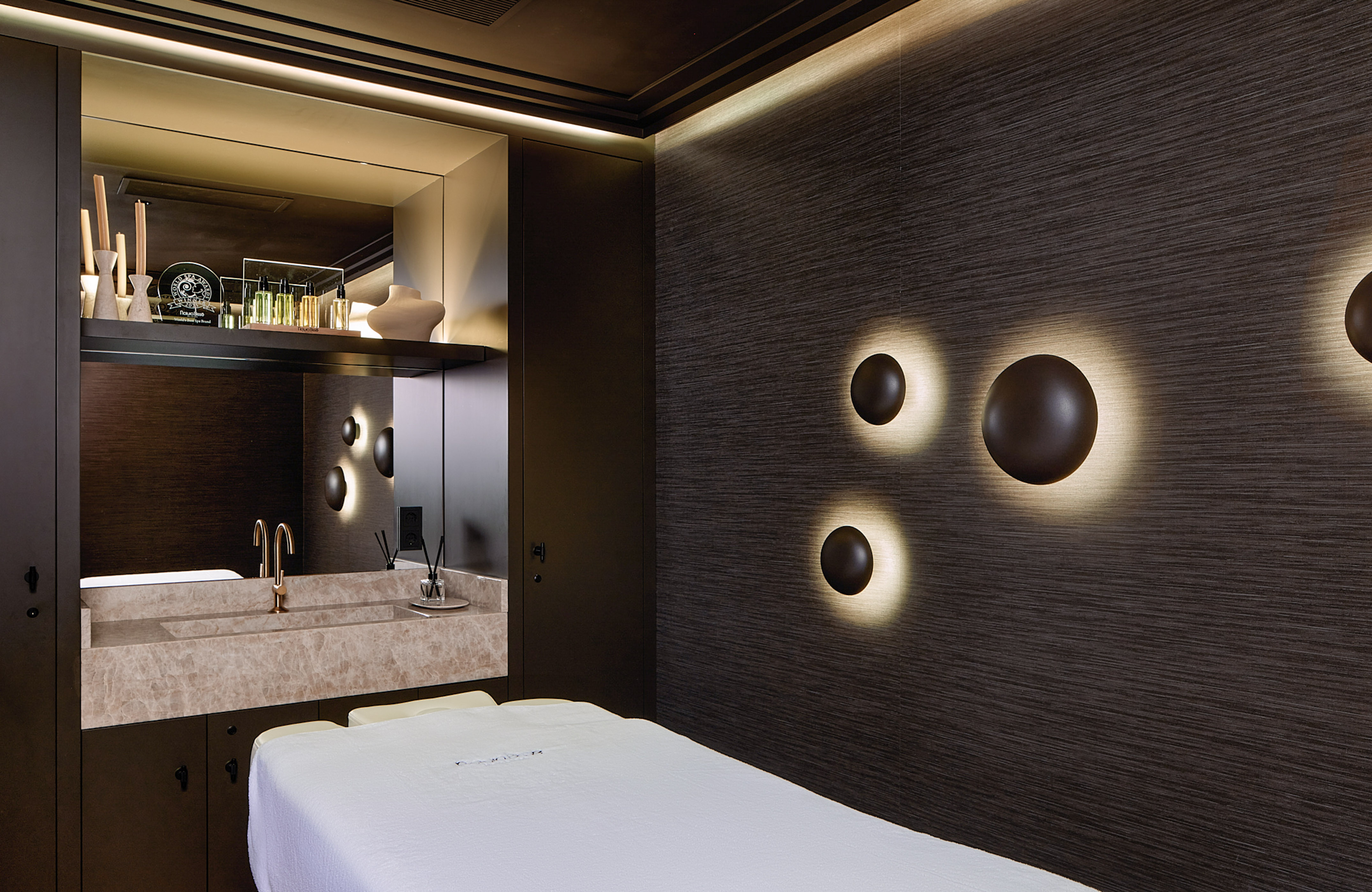 Dark luxury spa treatment room with sconce lights, marble vanity, and massage bed.