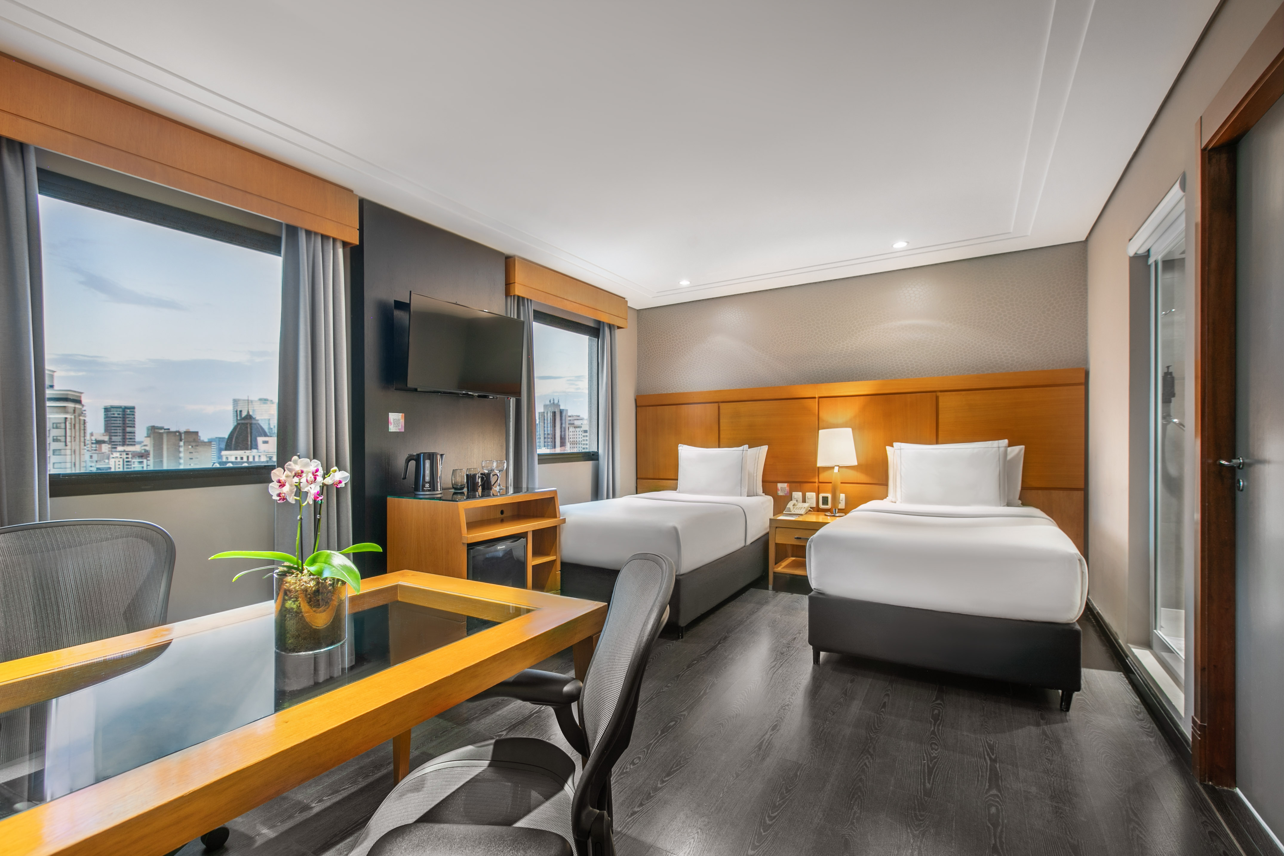 Modern hotel room with two twin beds, city view, desk, and TV.